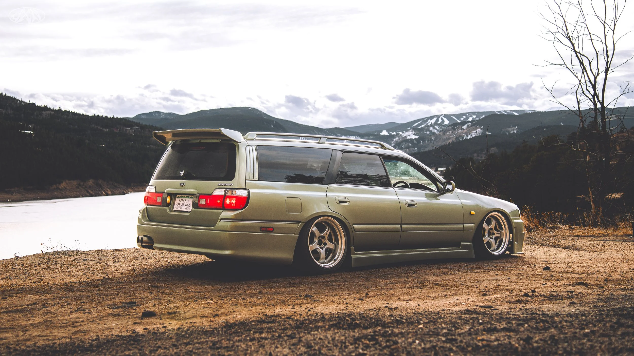 Green station wagon parked on a dirt area near a river with mountains and cloudy sky in the background.