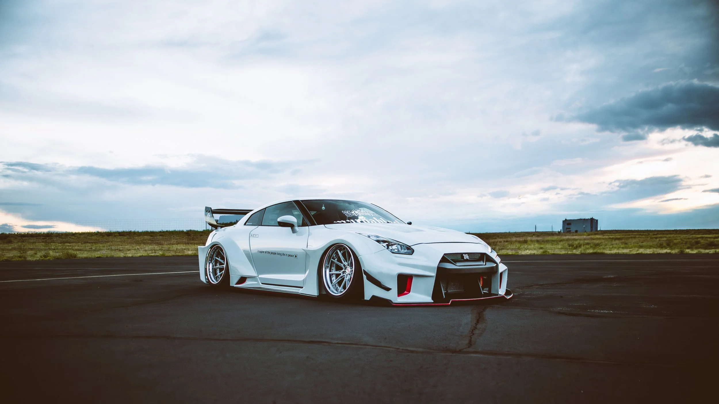White sports car with a large rear spoiler parked on an empty road in a rural setting under a cloudy sky.
