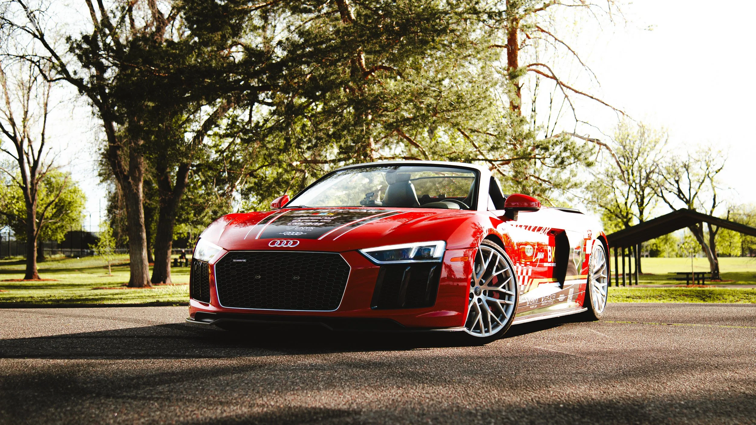 Red Audi sports car with racing decals parked on a paved area in a park with green grass, trees, and a pavilion in the background.