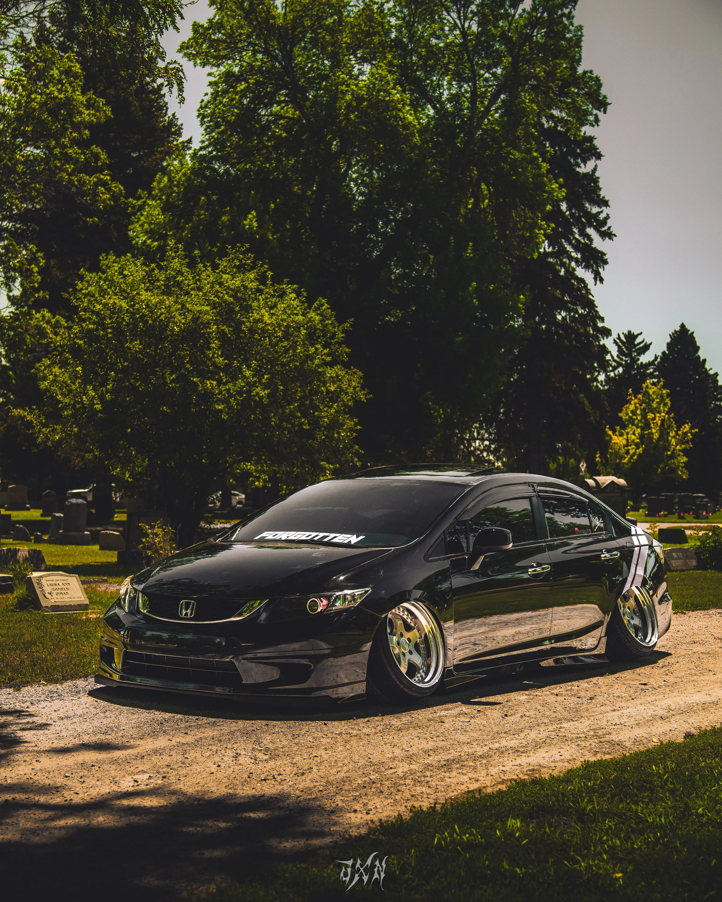 Black Honda Civic car with aftermarket wheels parked on a gravel path in a cemetery with tombstones and green trees in the background.
