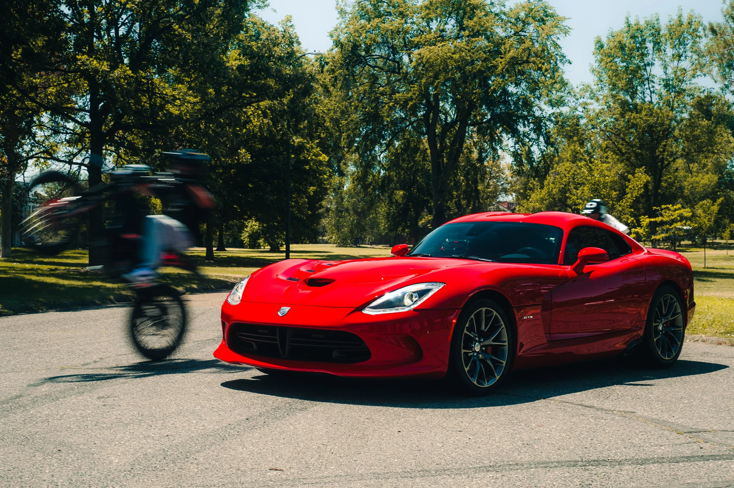 A red sports car parked on the side of a road with green trees and clear blue sky in the background. A cyclist riding past the car is blurred, indicating motion.