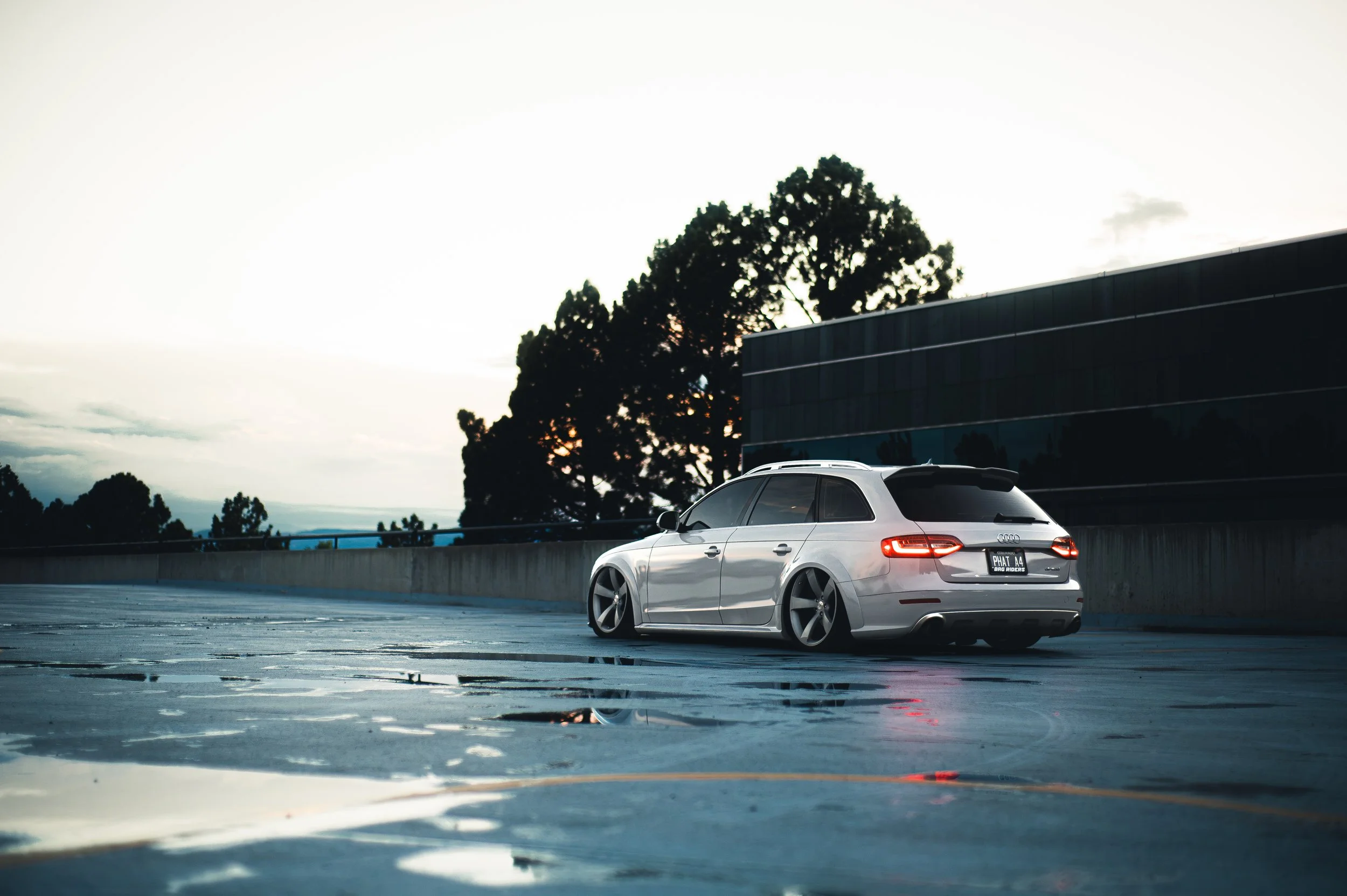 A silver Audi station wagon parked on a top level of a parking garage at sunset or early evening, with a backdrop of trees and a modern building with reflective glass windows.