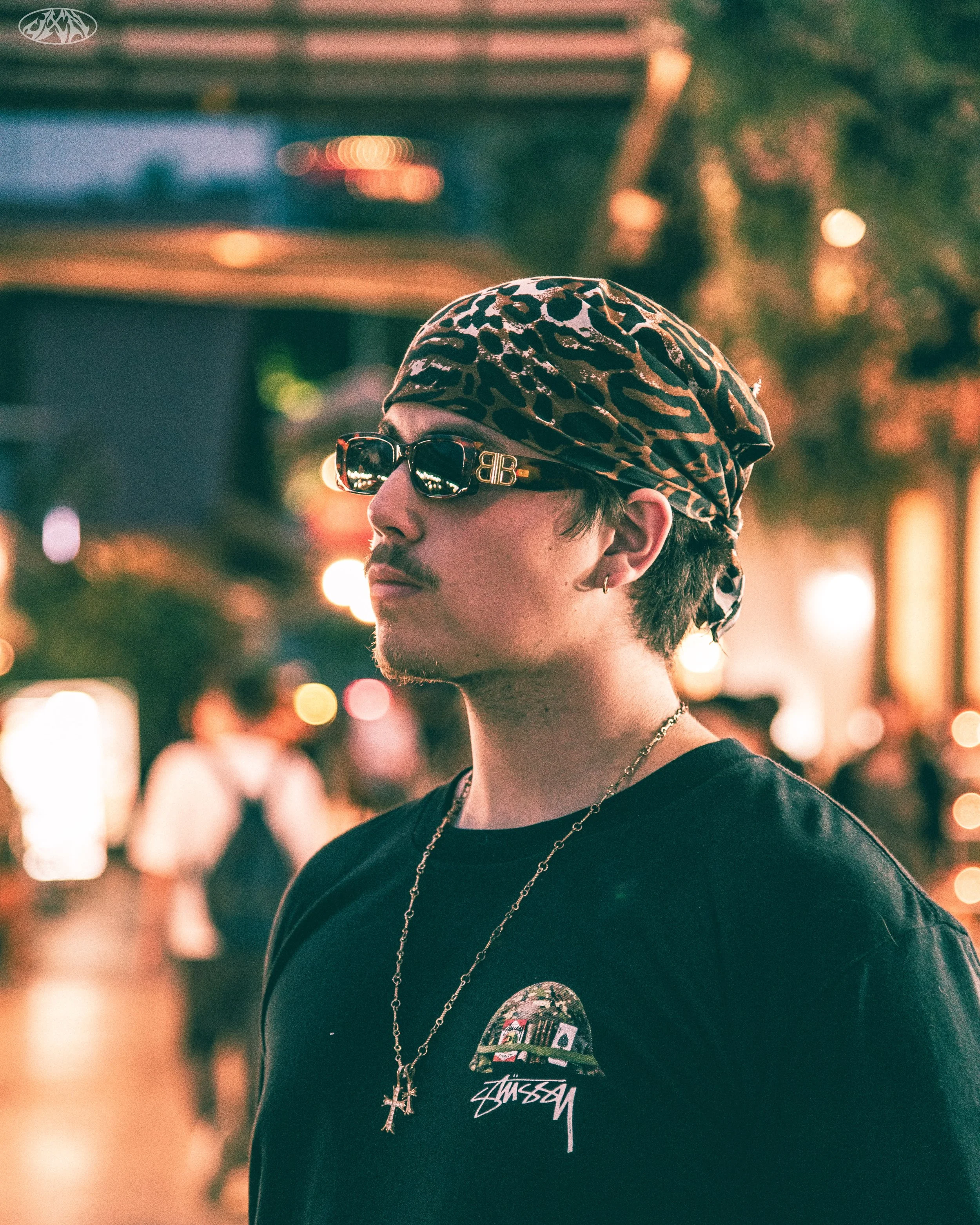 A young man with a leopard print headscarf and sunglasses, wearing a black T-shirt with a graphic and a gold chain, stands outdoors at night with blurred city lights and people in the background.