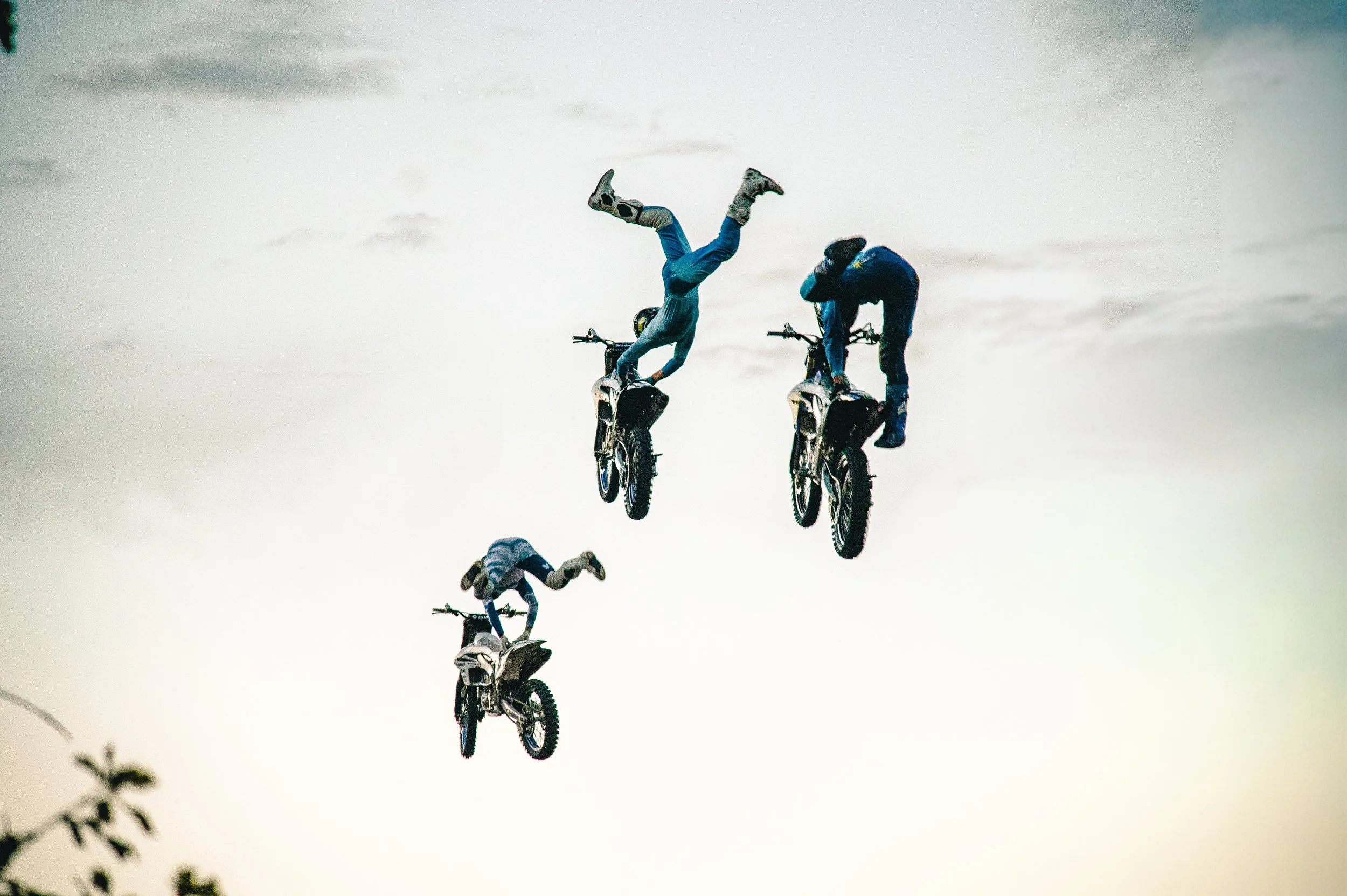 Four bikers perform mid-air stunts against a cloudy sky, with one biker executing a handstand on the handlebars.