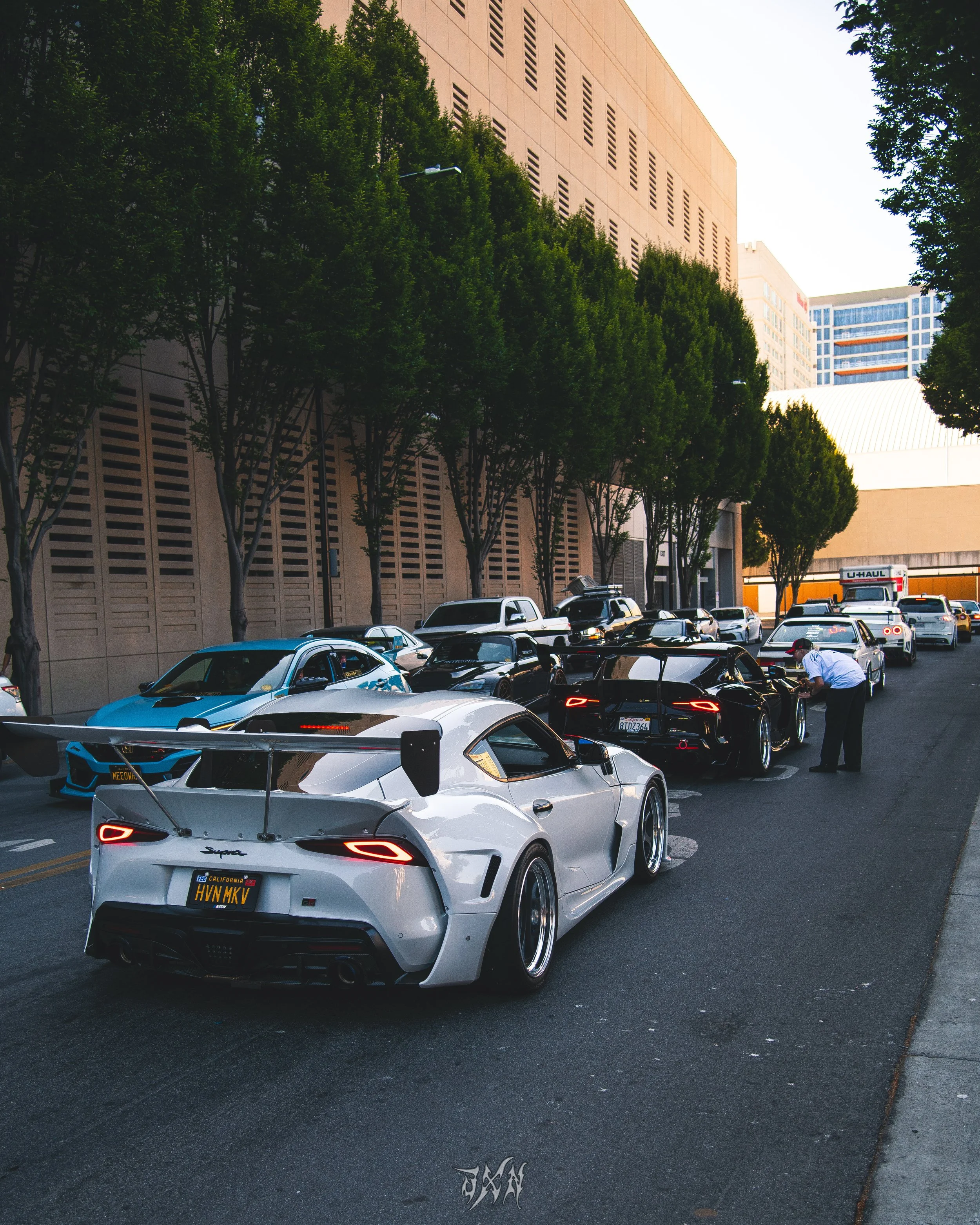 A white sports car with a large rear wing, parked among other cars on a city street with tall buildings and green trees.