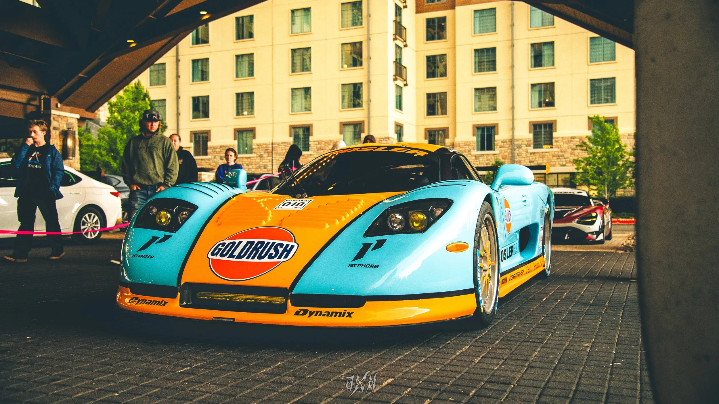 A bright blue and orange race car with sponsor logos, parked under a bridge with people standing around it, and a hotel building in the background.