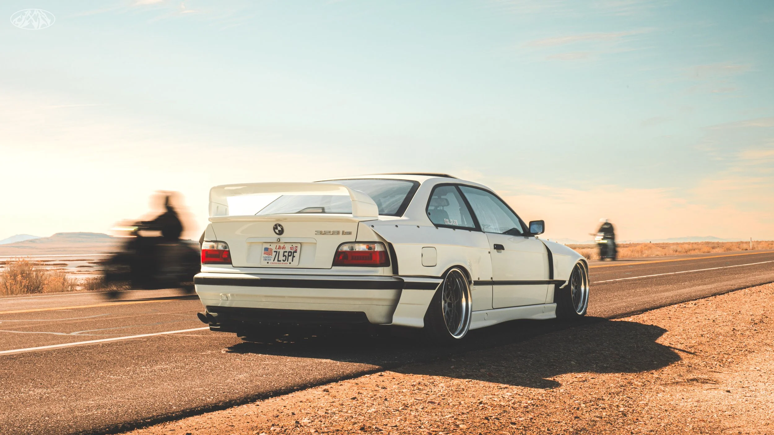 A white BMW 3 Series car with a large rear spoiler parked on the side of a highway. Motion-blurred motorcycles are driving past. The landscape is open with desert-like terrain and a mountain range in the distance under a soft, partly cloudy sky.