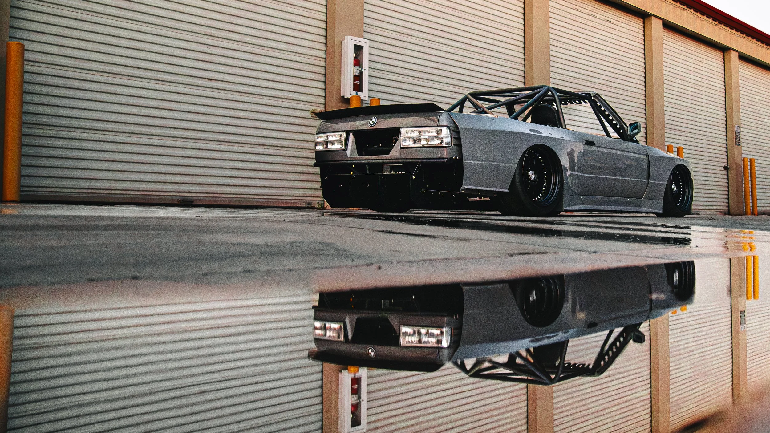 A gray race car with a black roll cage parked inside a garage with closed roll-up doors and a fire alarm box on the wall.
