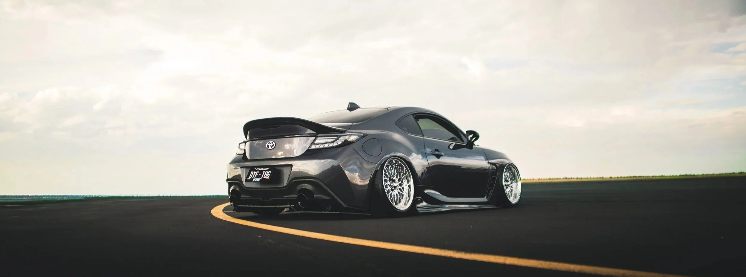 A black sports car with custom wheels, low profile, and a spoiler on the rear, parked on a curved asphalt road under a cloudy sky.