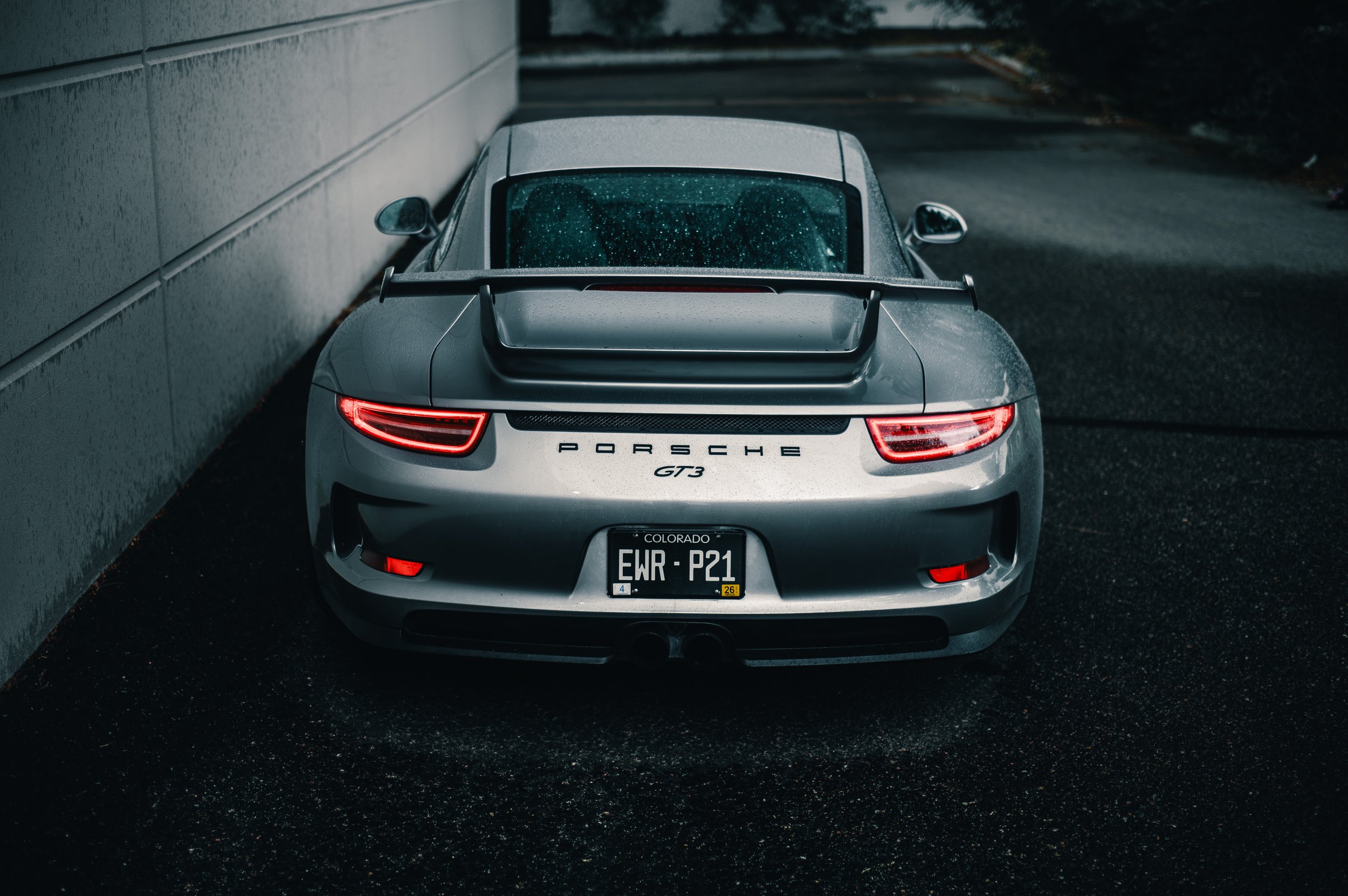A silver Porsche GT3 car parked on a wet pavement next to a grey building, with rain droplets visible on the rear windshield and taillights turned on.