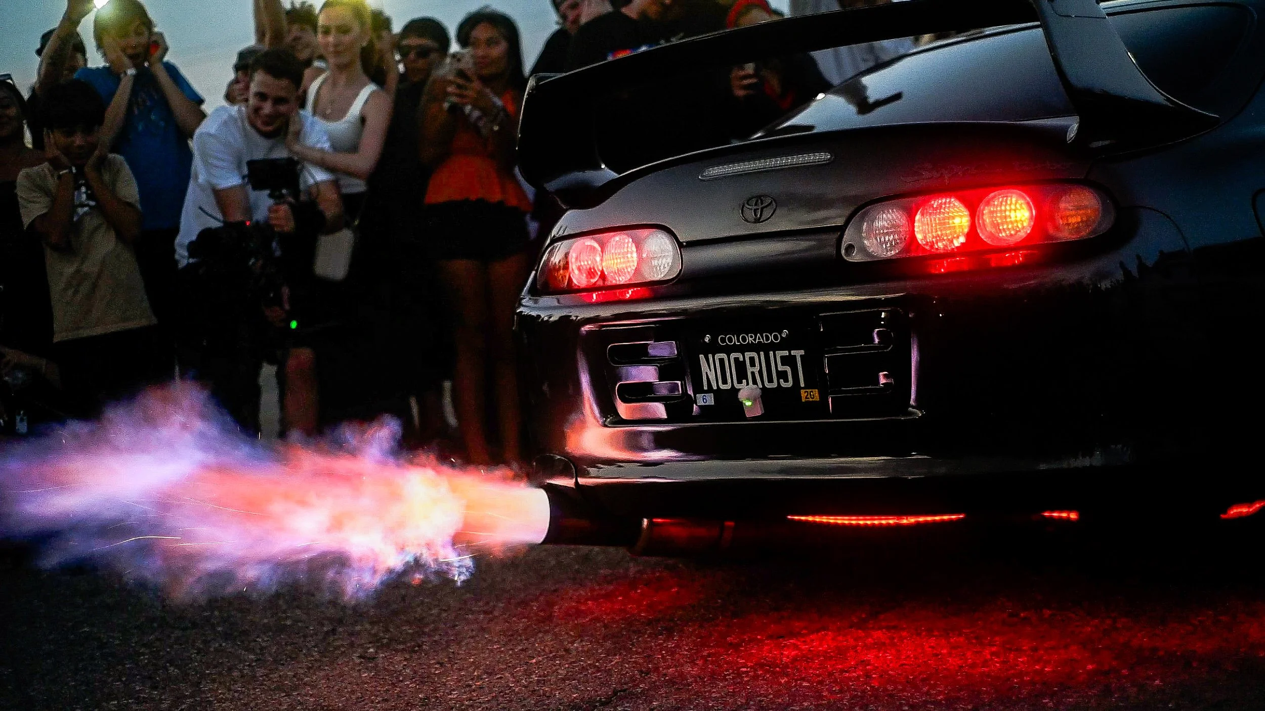 A black Toyota car with a Colorado license plate that reads 'NOCRUST' is performing burnout, emitting flames from the exhaust, during an event with a crowd of people watching and taking photos.