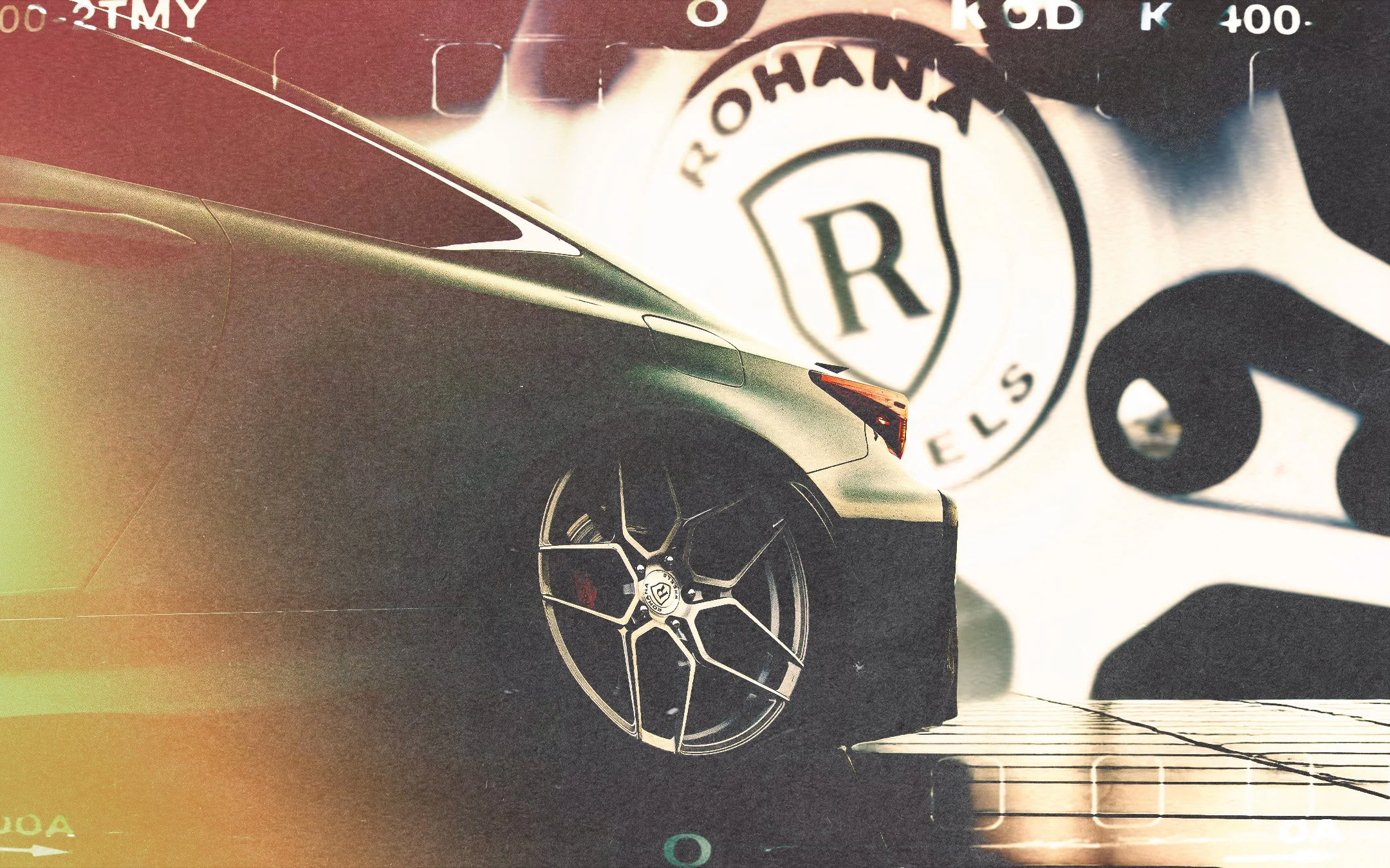 Close-up view of a silver car's front wheel and part of its body, with a blurred RBI logo sticker on the top right corner.