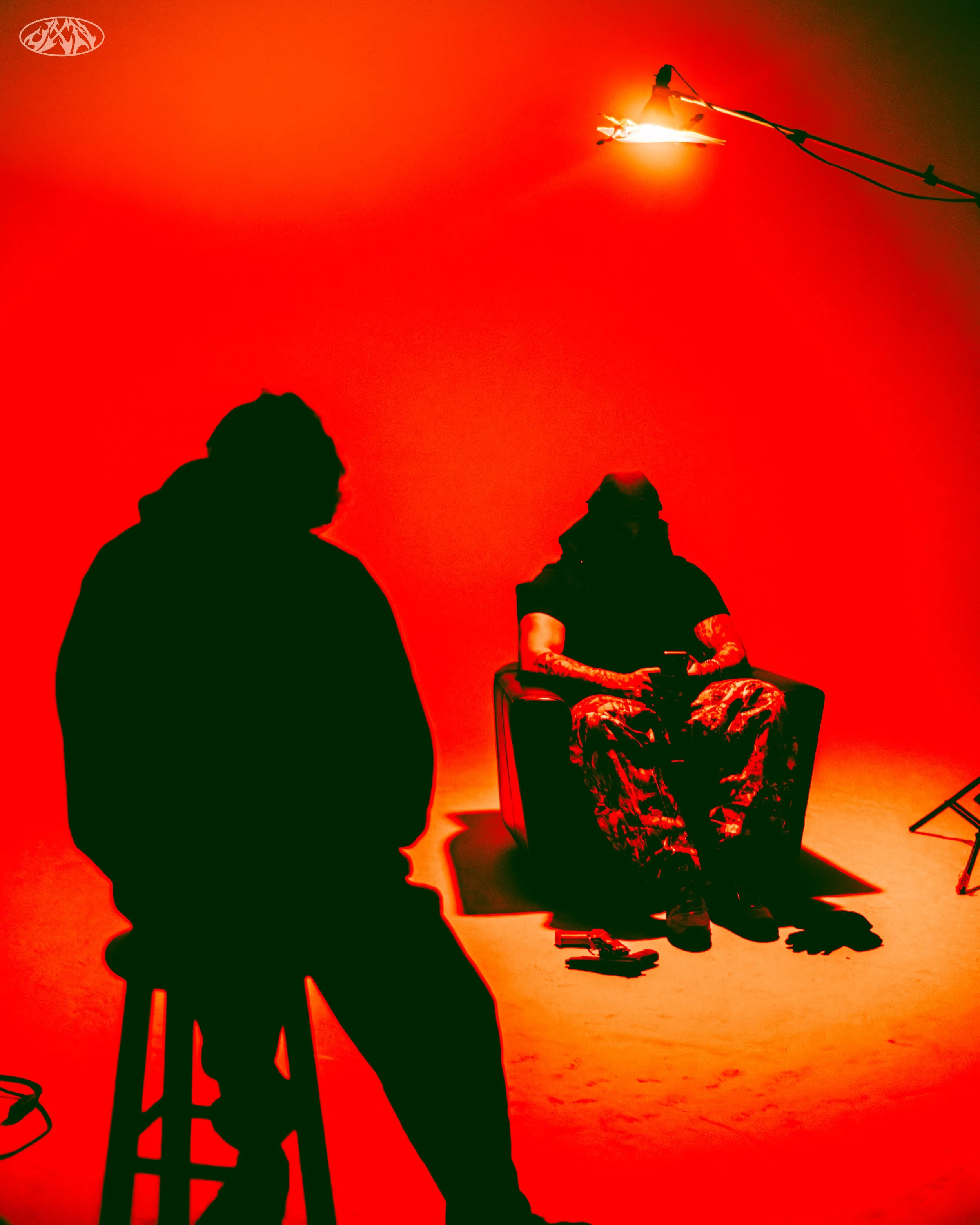 Silhouettes of two people in a dark room with a red background, one sitting on a stool and the other using a laptop, with a professional lighting setup overhead.