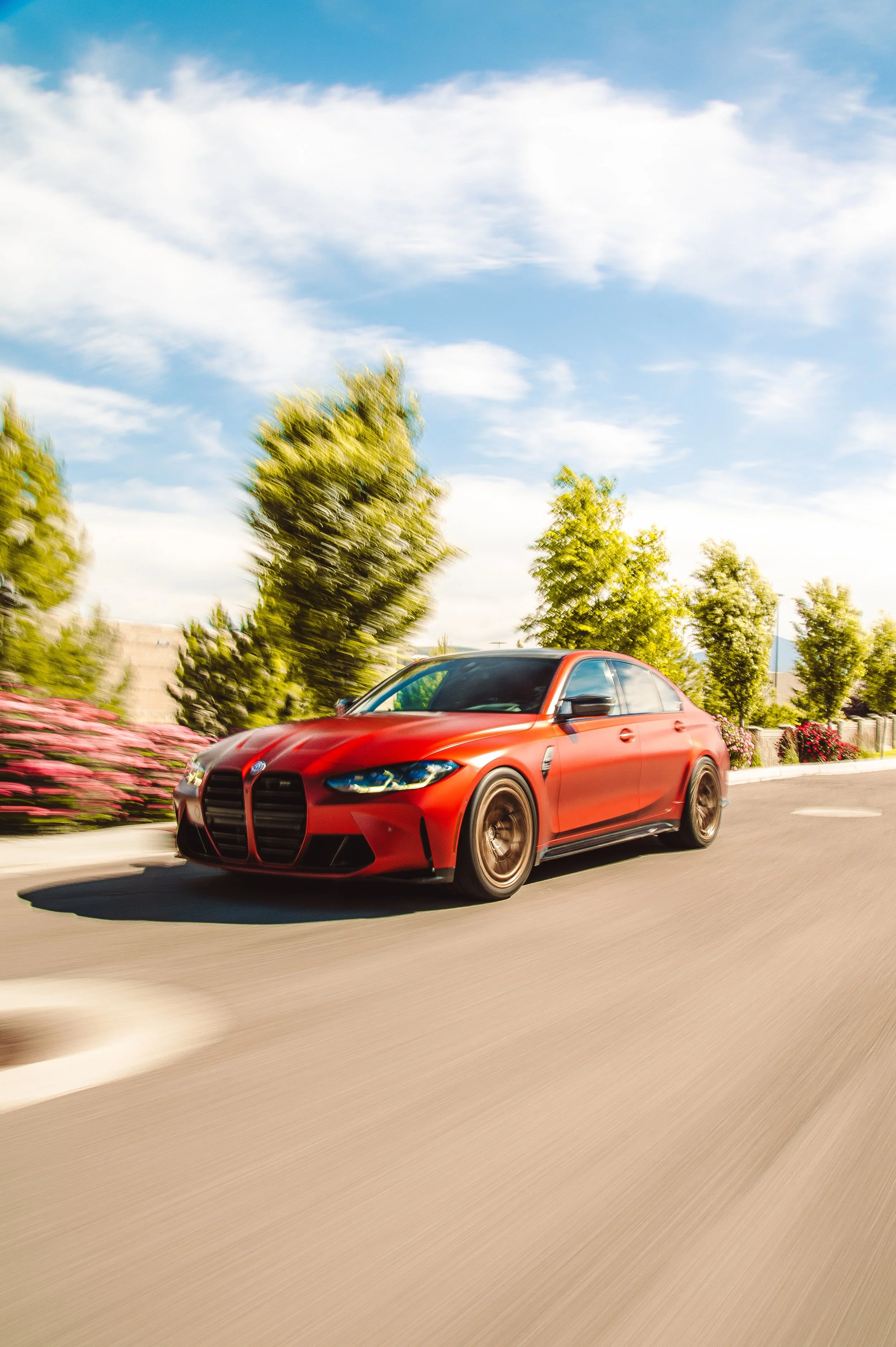 A red sports car driving on a road with trees and pink flowers on the side, under a partly cloudy sky.