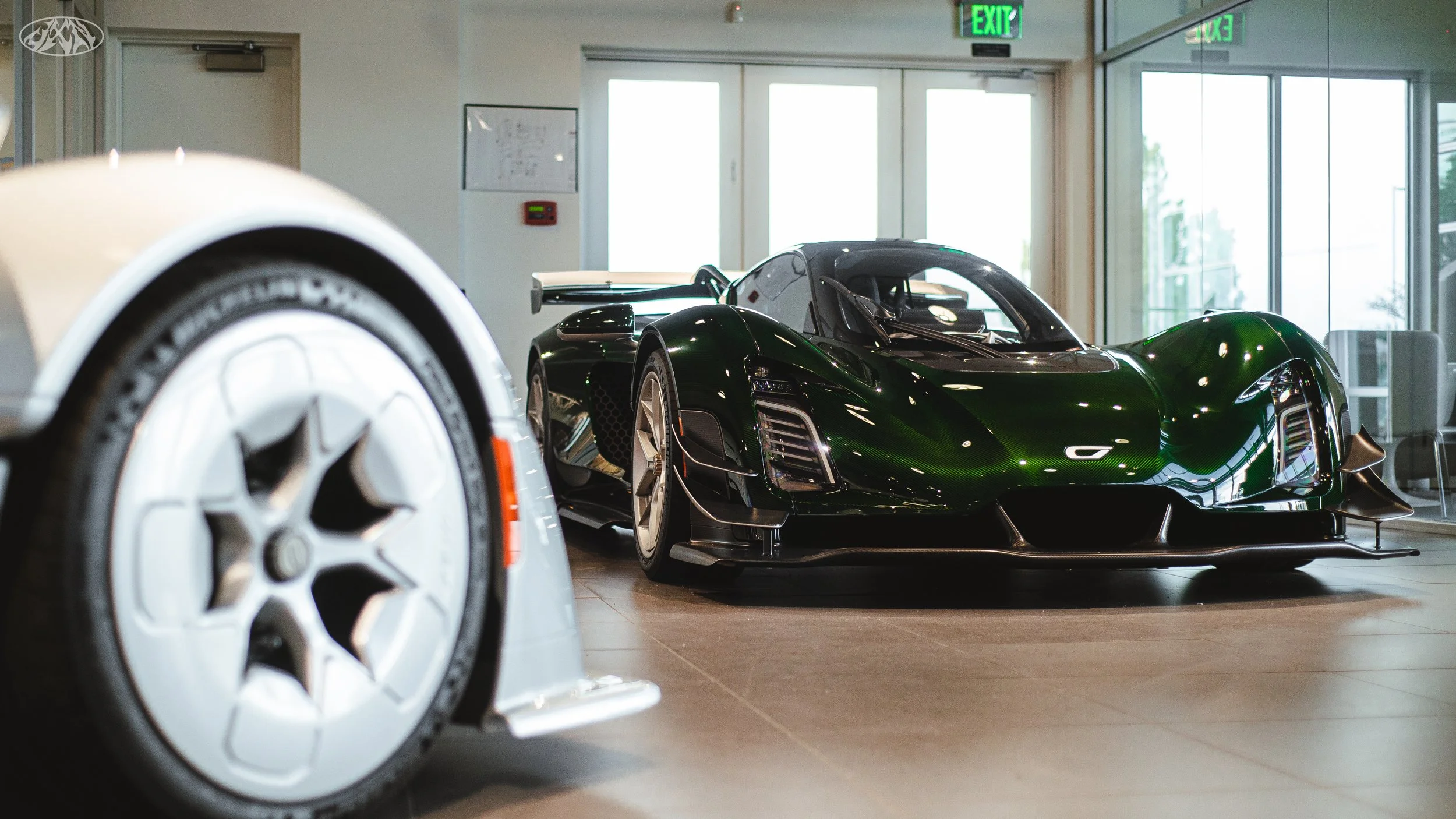 A black racing sports car with a large rear wing on display inside a showroom or garage, with another vehicle partially visible in the foreground.