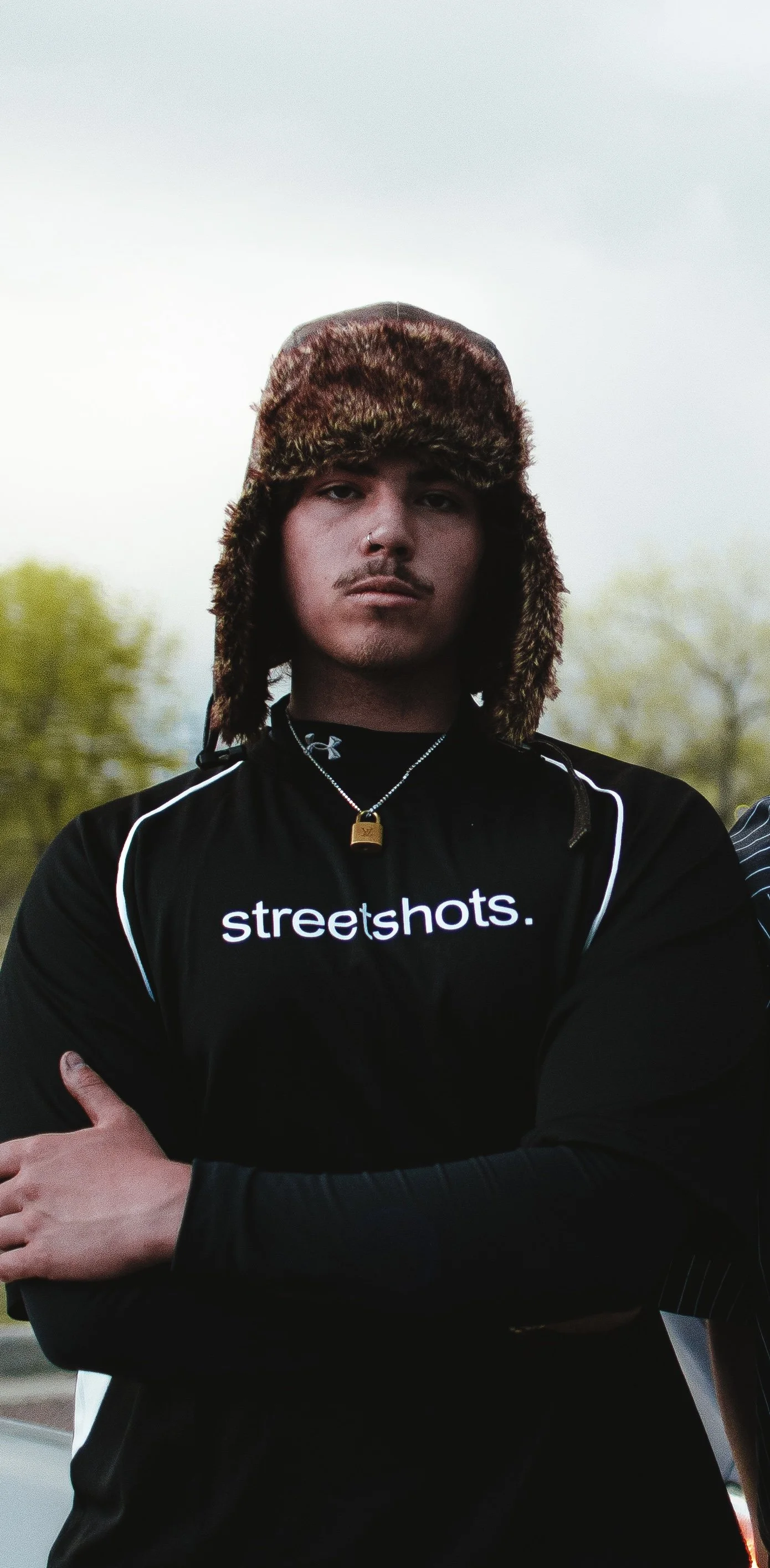 A young man wearing a furry winter hat, black sports shirt with white writing that says 'streetshots.', and a silver chain with a padlock pendant, stands outdoors with arms crossed, trees blurred in the background.