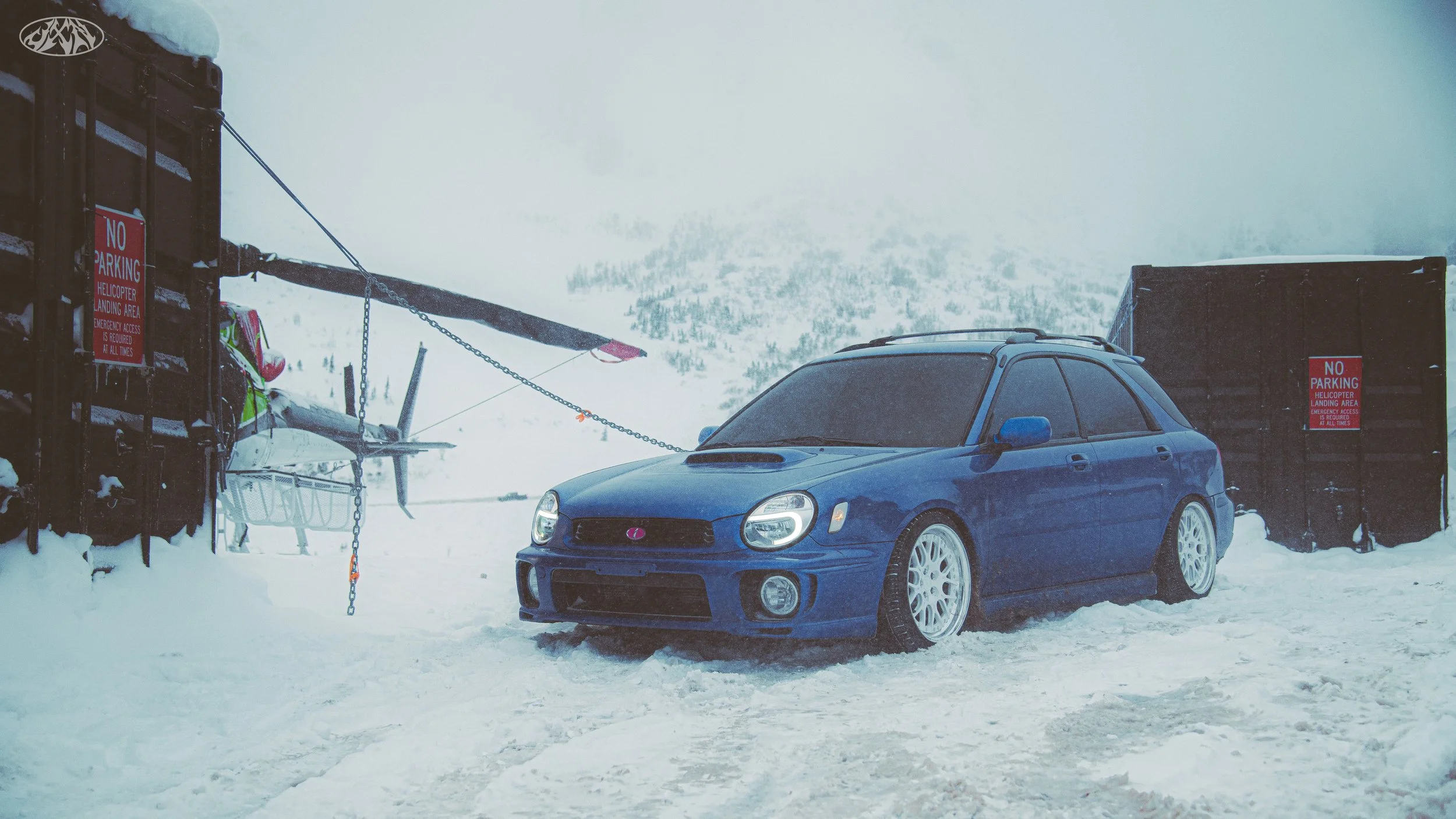 A blue Subaru car parked in a snowy area with ski equipment and a sign that says 'No Parking'. There is a mountain landscape in the background, partially obscured by fog or clouds.