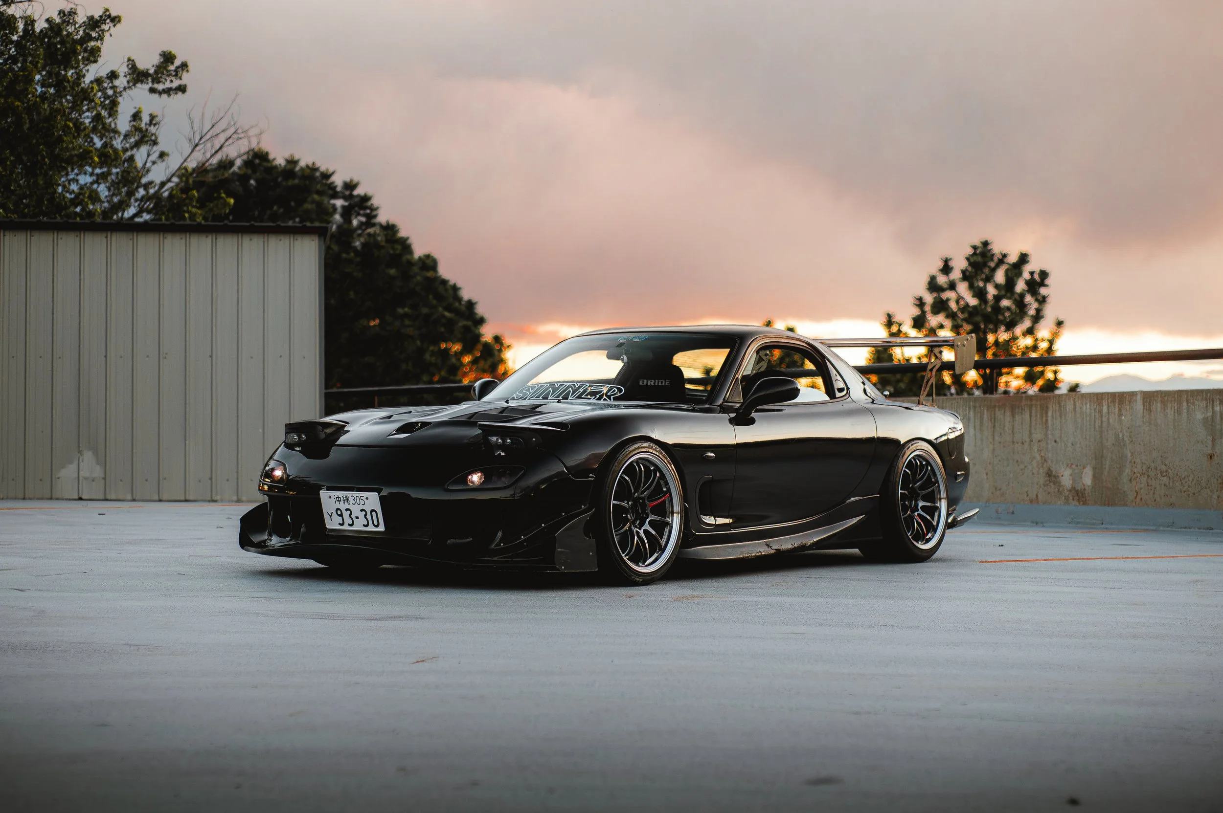 A black sports car with modifications, including a large rear spoiler, is parked on an empty rooftop parking lot during sunset. The background includes trees and a cloudy sky.