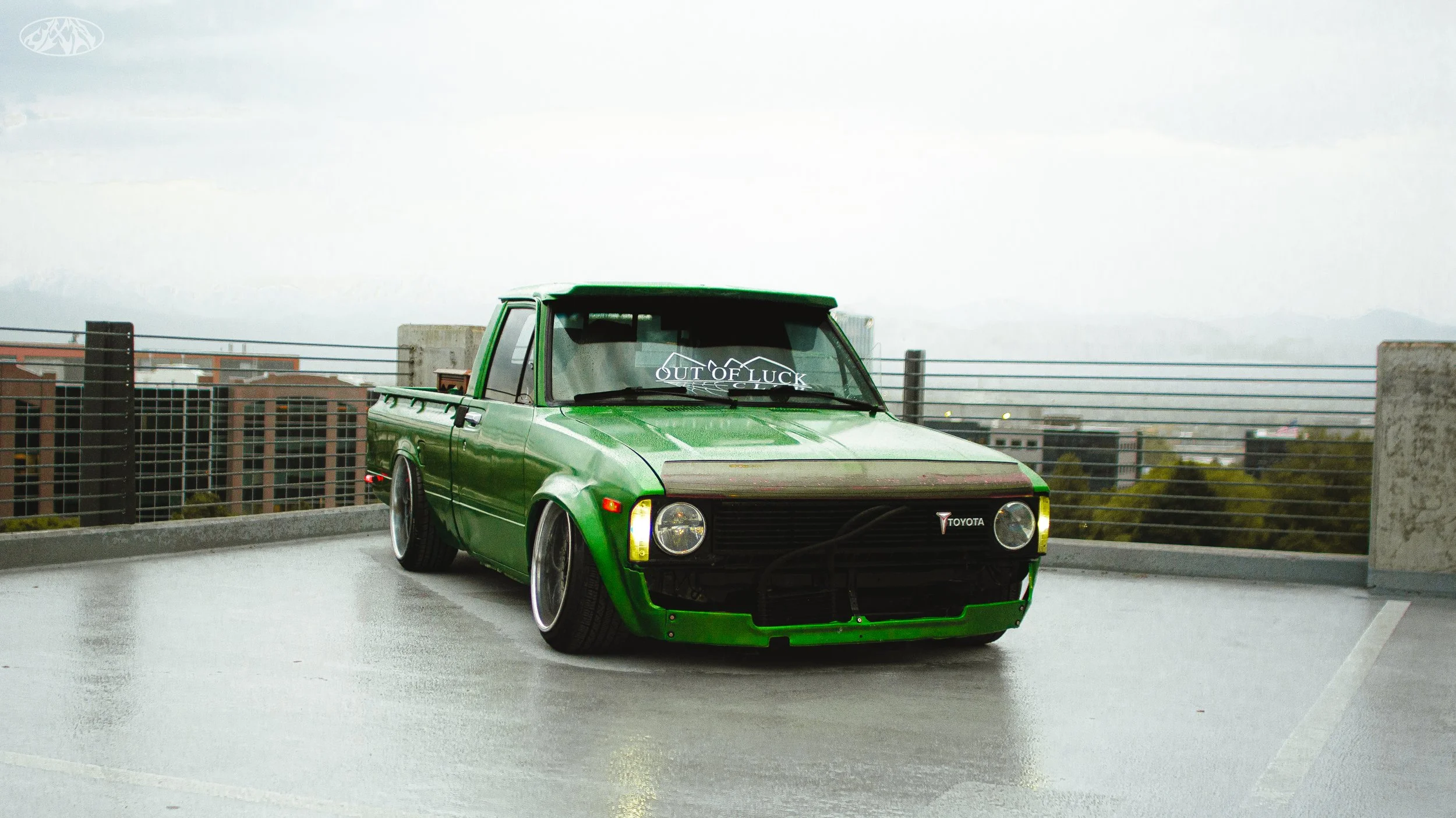 Green modified Toyota pickup truck parked on a rooftop parking lot with city buildings and mountains in the background.