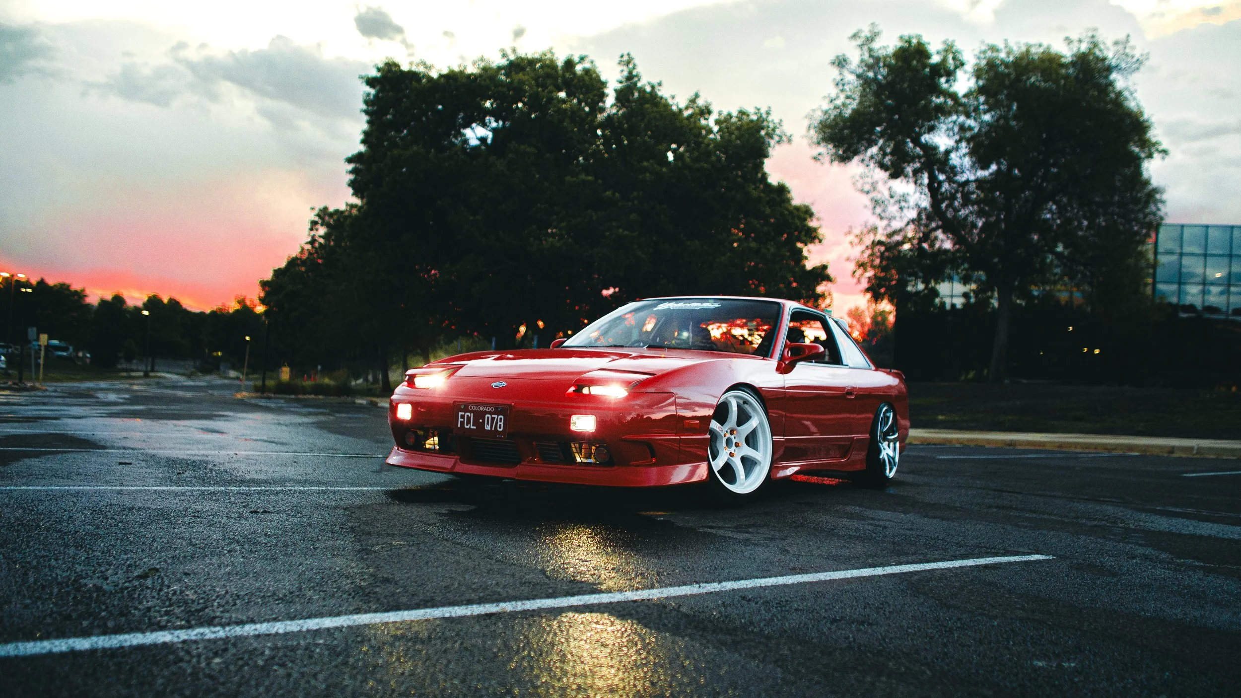 A red sports car parked on a wet parking lot during sunset, with trees and a glass building in the background.