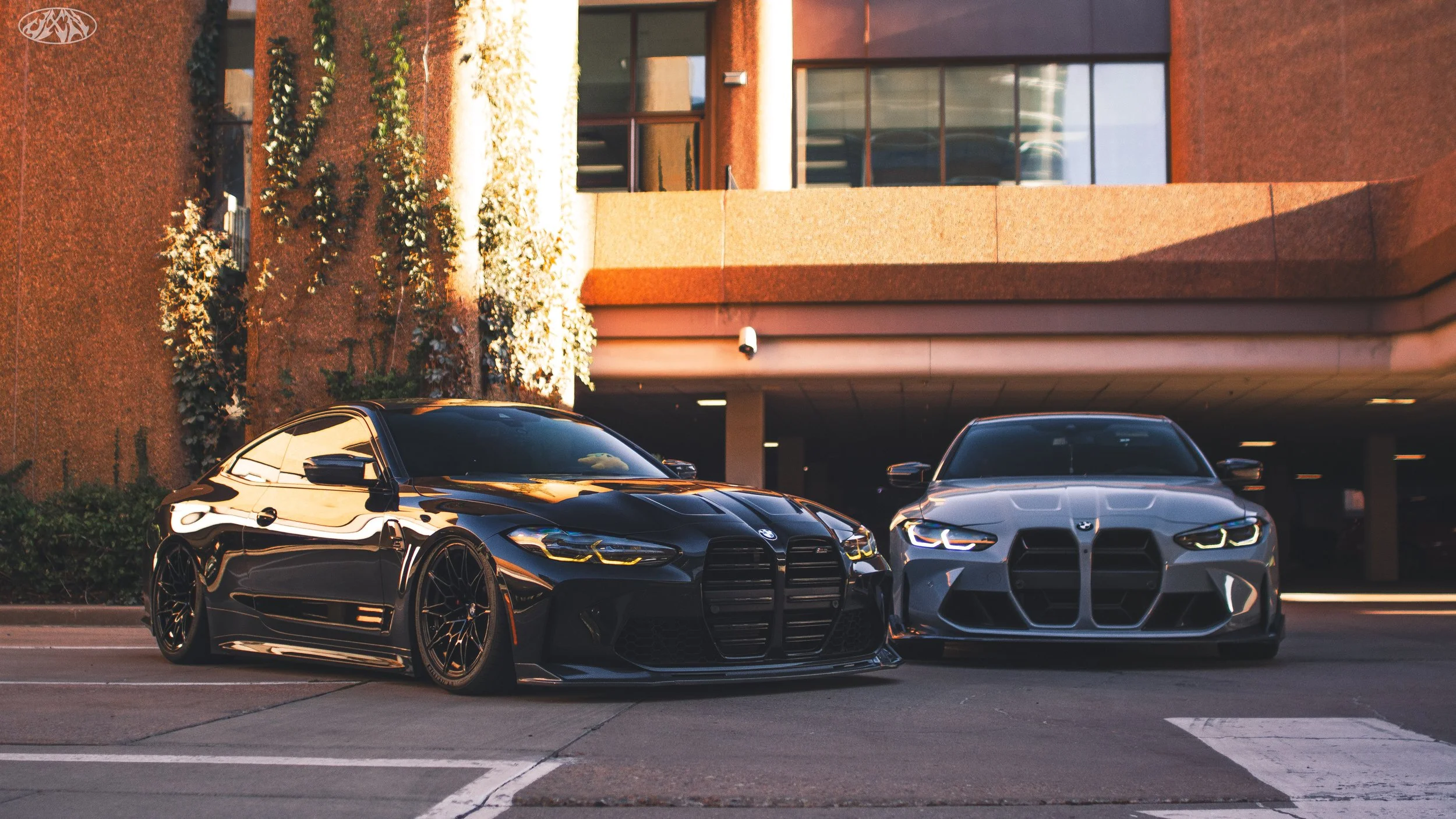 Two luxury cars, a black car and a silver car, parked in front of a modern building with large windows and a brown exterior, with some bushes and flowering plants nearby, during sunset.