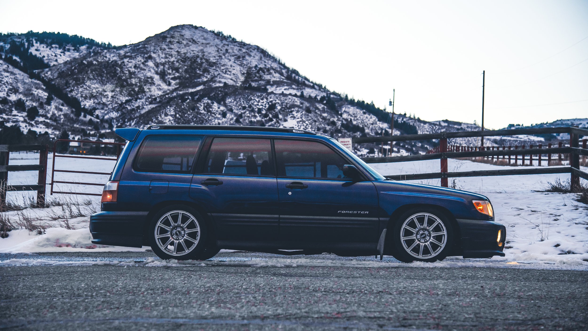A blue Subaru Forester parked on the side of a snow-covered road with a mountain in the background.