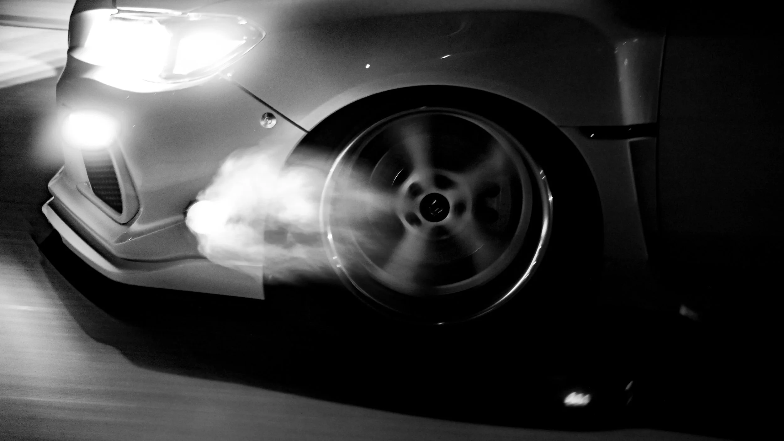 A black and white photo of a sports car in motion, showing the front left side and wheel, with smoke coming from the tire.