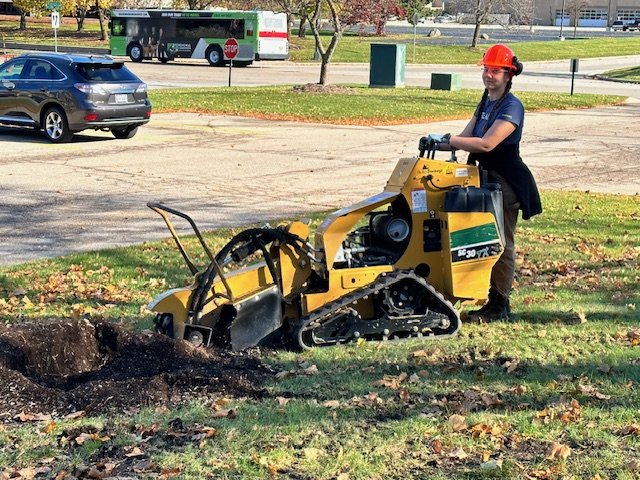 Woman wearing a red helmet operating a small yellow trench compactor machine on grass with a hole in the ground. Background includes parked cars, a bus, and trees.