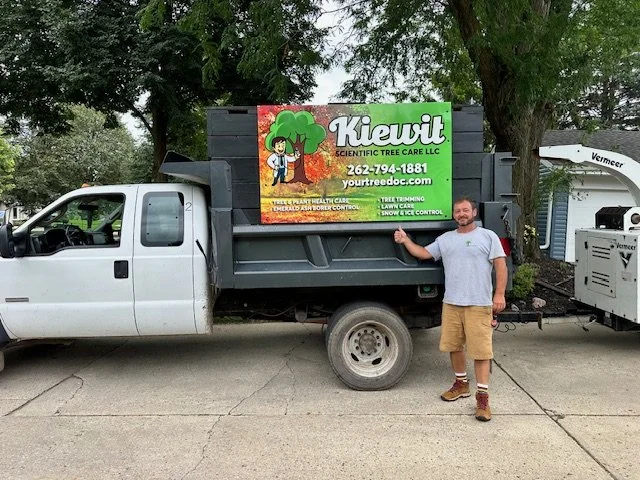 Man standing next to a white truck with a sign for Kieuit Scientific Tree Care LLC on the bed, tree care services displayed.