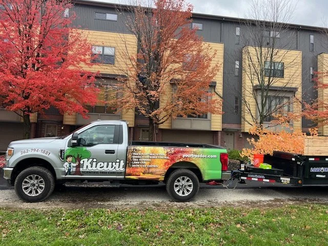 A pickup truck with the text "Kiewit" and a dinosaur logo, parked in front of a building with autumn trees. There is a trailer attached to the truck.