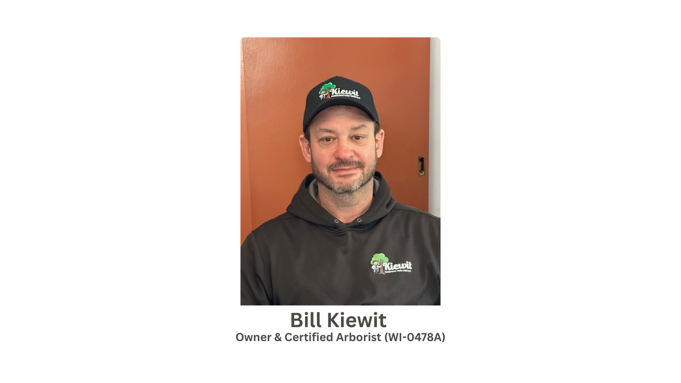 Photograph of Bill Kiewit, a man with a beard, wearing a black cap and black hoodie, both with a logo and text related to Kiewit. The background is a mix of orange-brown and white walls. A caption below identifies him as the owner and certified arborist.