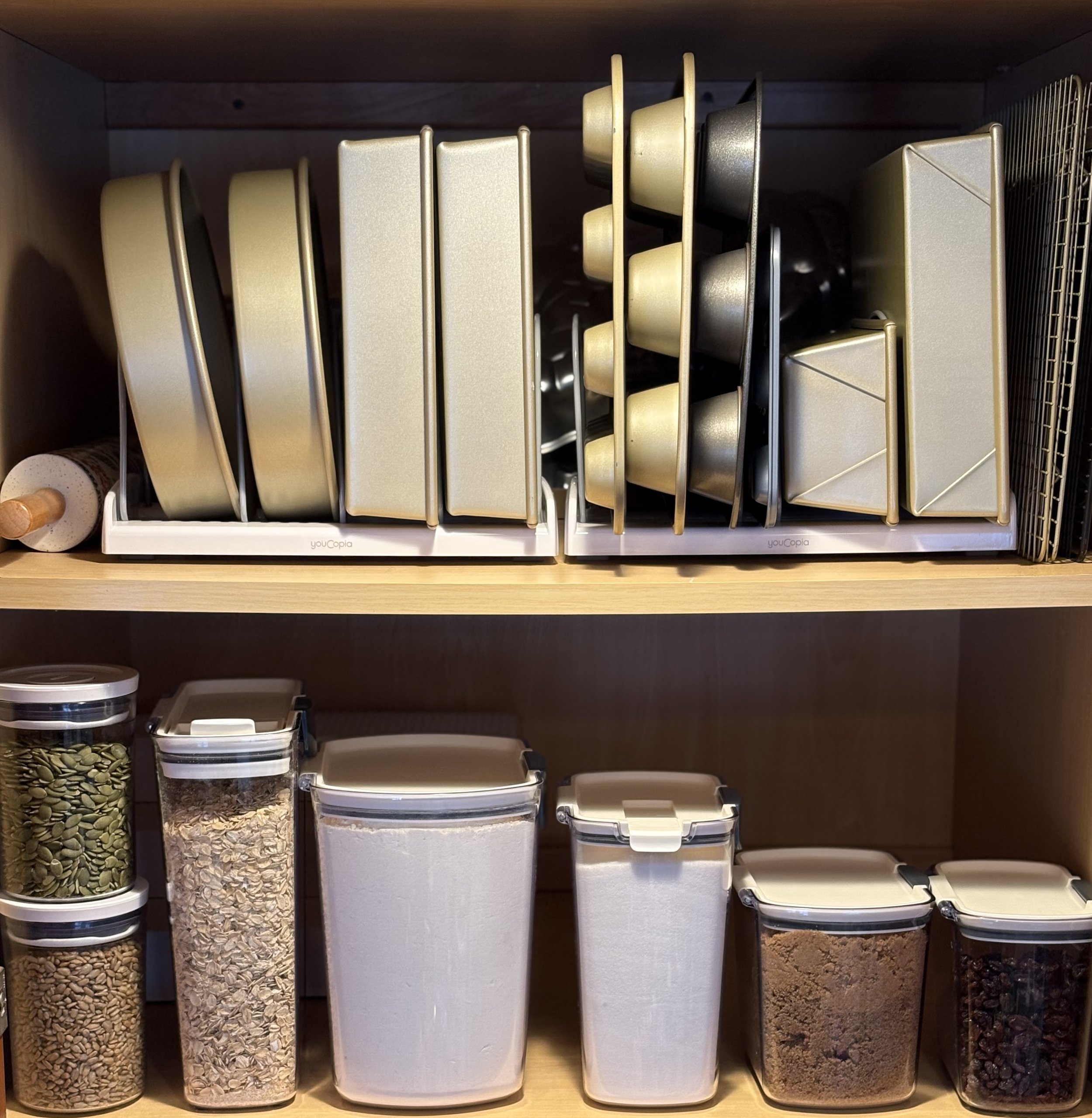Kitchen storage with beige and silver baking pans on dish racks and plastic containers holding seeds, oats, flour, and spices on a wooden shelf.