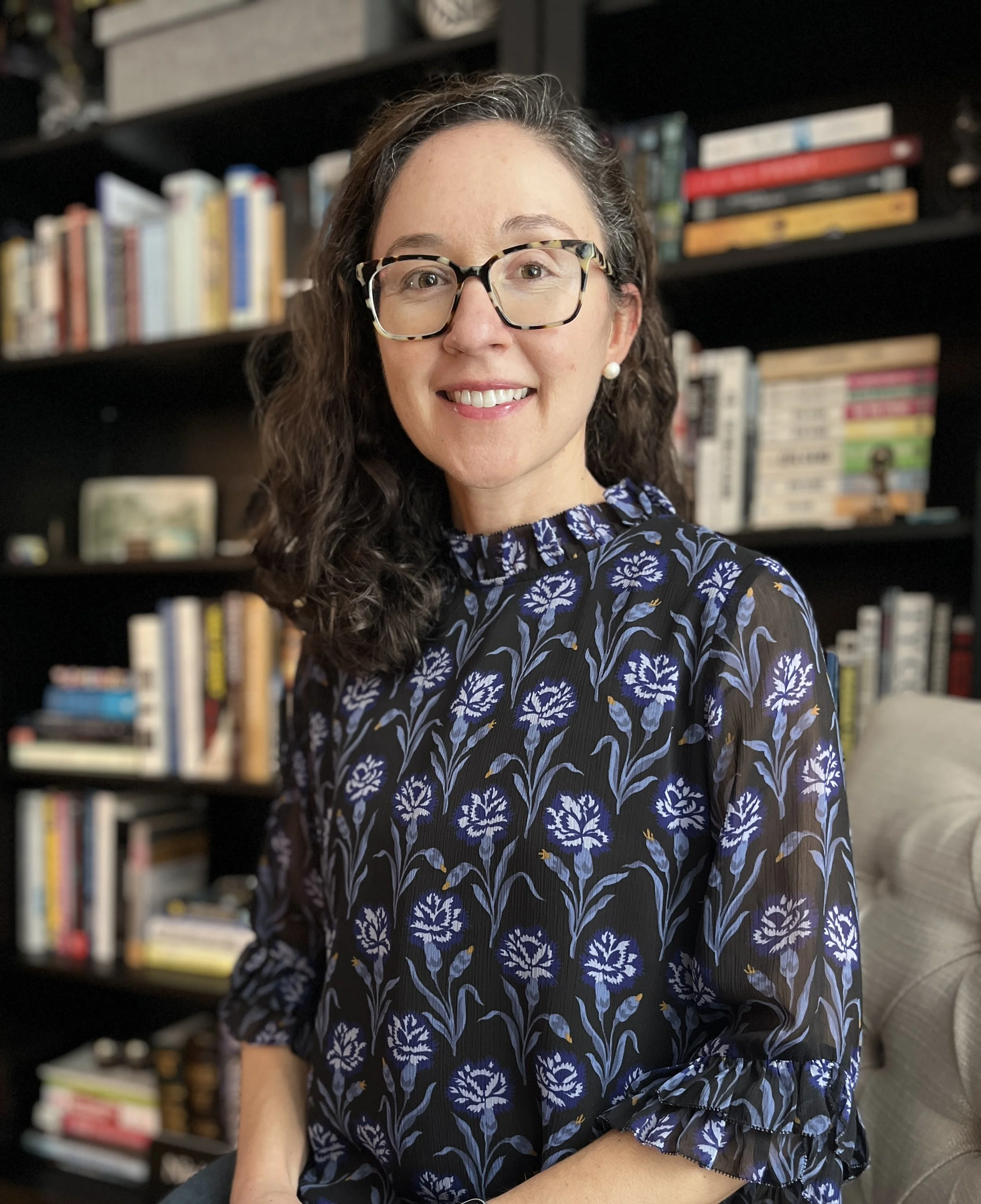 A woman with curly dark hair, glasses, and pearl earrings, smiling while sitting in a room with a bookshelf filled with books behind her. She is wearing a dark blouse with a blue and purple floral pattern.