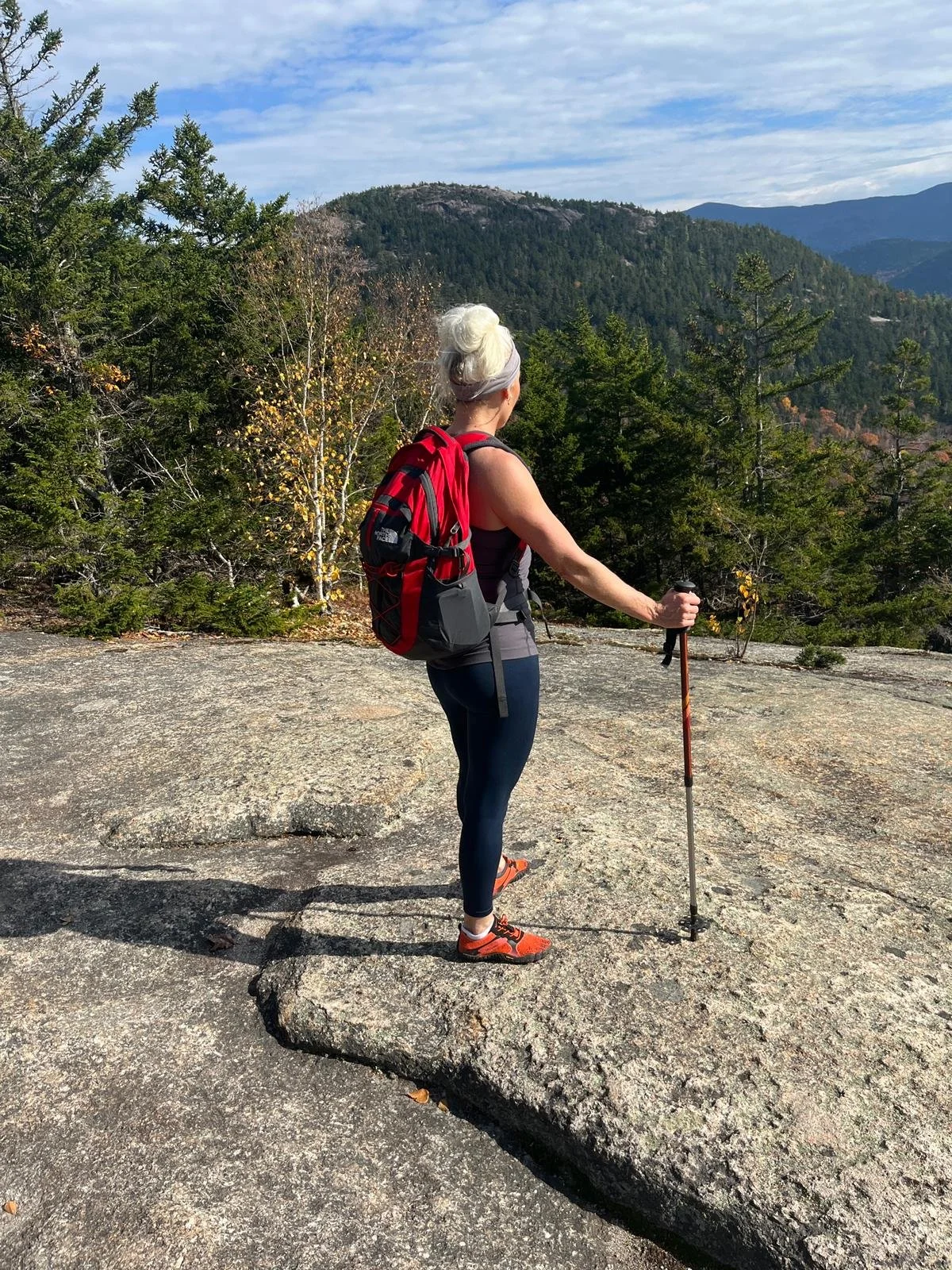 A woman with white hair, wearing a black tank top, black leggings, and orange hiking shoes, stands on a large rock with a hiking pole, overlooking a forested mountain landscape during daytime.