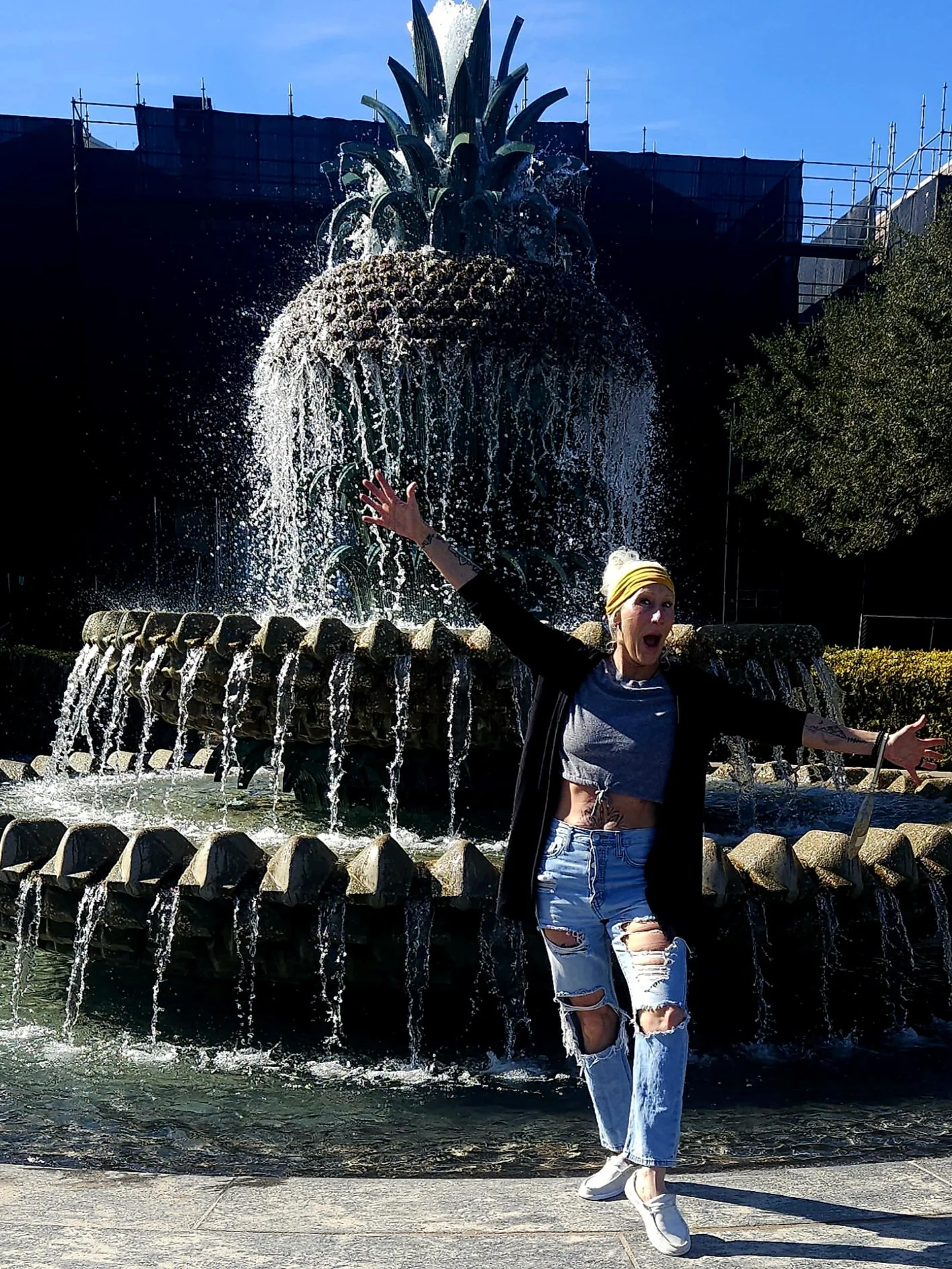 A woman posing enthusiastically in front of a pineapple-shaped fountain during daytime, splashing water and stretching her arms wide.
