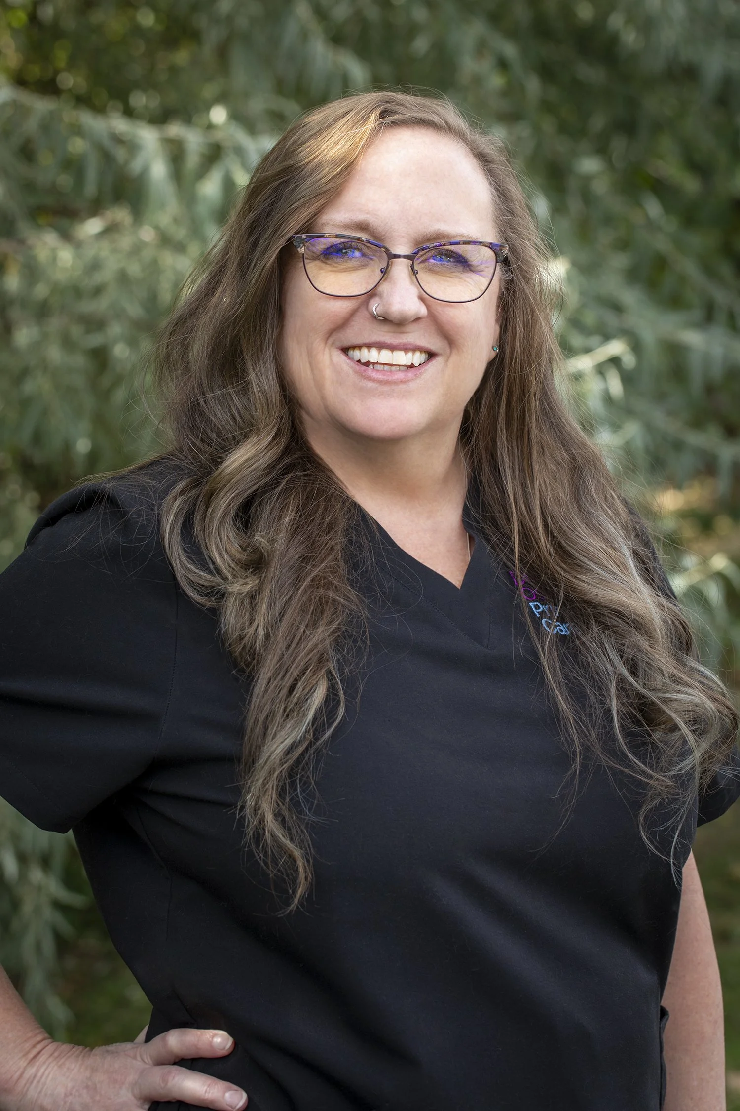 A woman with long wavy brown hair, glasses, a nose ring, and teal earrings, wearing a black shirt, standing outdoors with green foliage in the background.