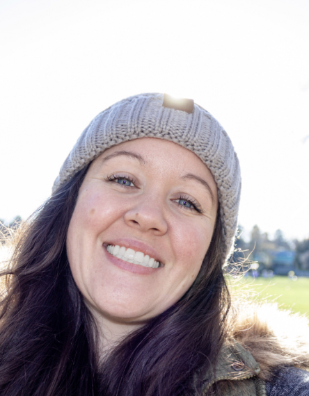 Close-up of a smiling woman wearing a gray knit beanie outdoors on a sunny day.