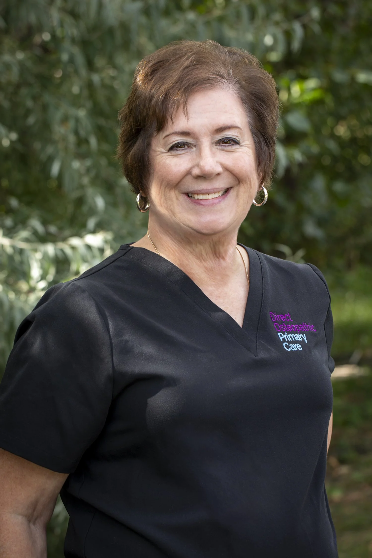 A woman smiling outdoors, wearing a black shirt with embroidered text reading "Direct Osteopathic Primary Care."