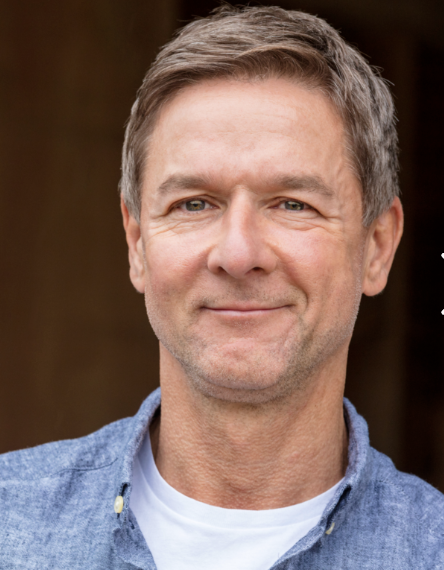Close-up of a middle-aged man with short gray hair, smiling, wearing a blue denim shirt over a white t-shirt, outdoors with a blurred background.