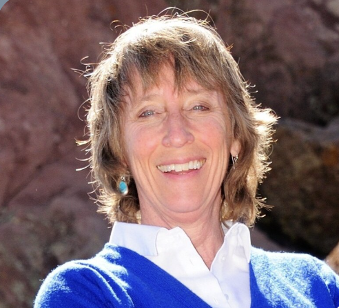A smiling woman with short, light brown hair wearing a blue sweater and white collared shirt outdoors.
