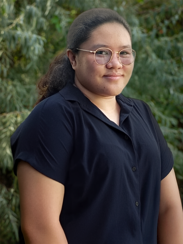 A woman with glasses and dark hair pulled back, wearing a navy blue shirt, standing outdoors with green foliage in the background.