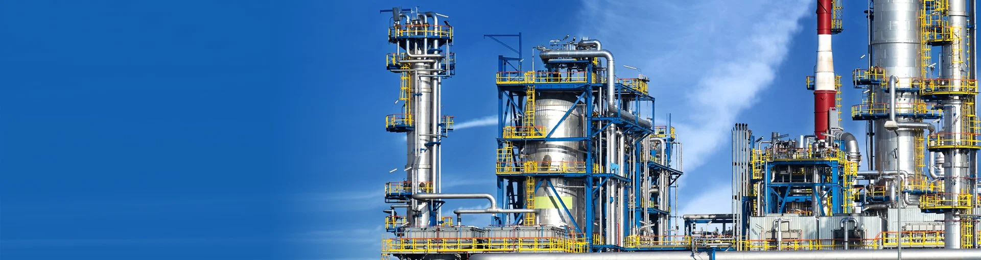 An industrial oil or chemical refinery with multiple large metal structures, pipes, and yellow safety railings against a clear blue sky.