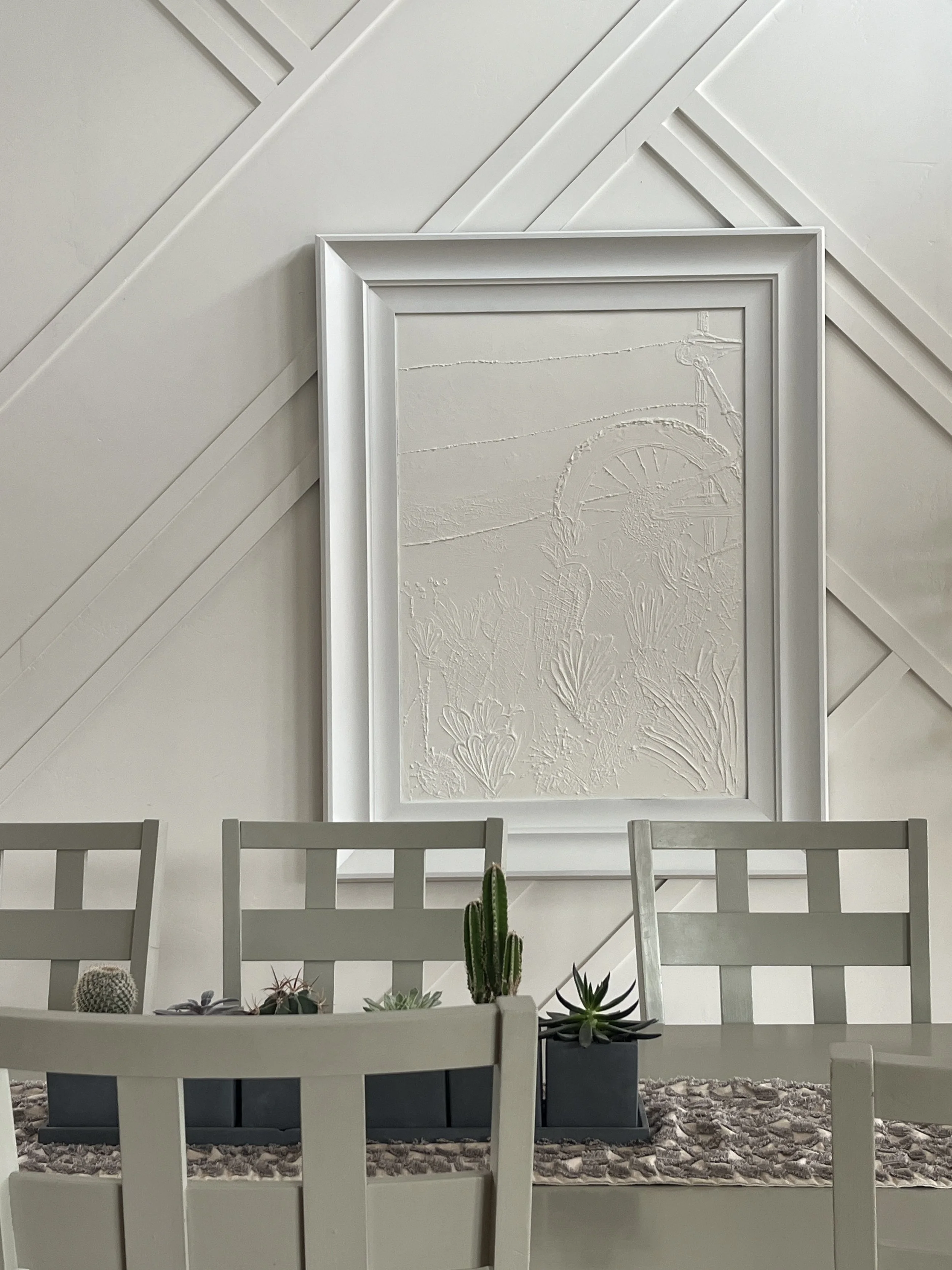 A white dining table with several white chairs, decorated with potted cacti and succulents, in front of a textured white wall with an abstract framed art piece.