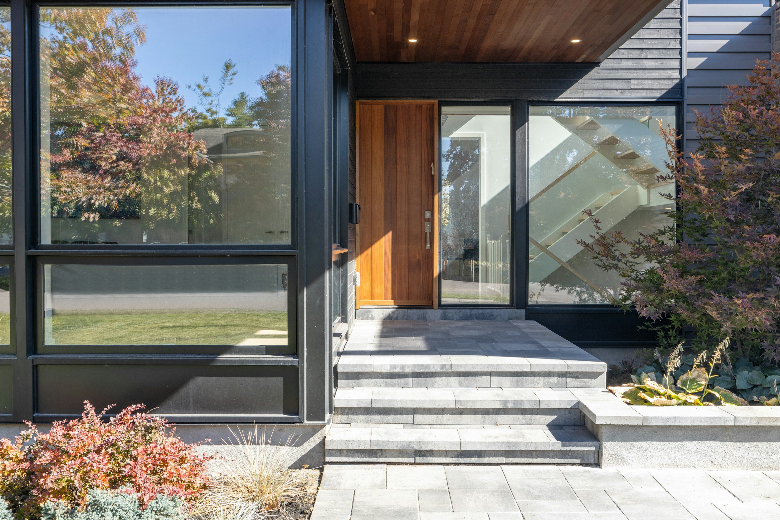 Modern house entrance with black exterior walls, wooden door, glass panels, stairs, and landscaping with plants and trees.