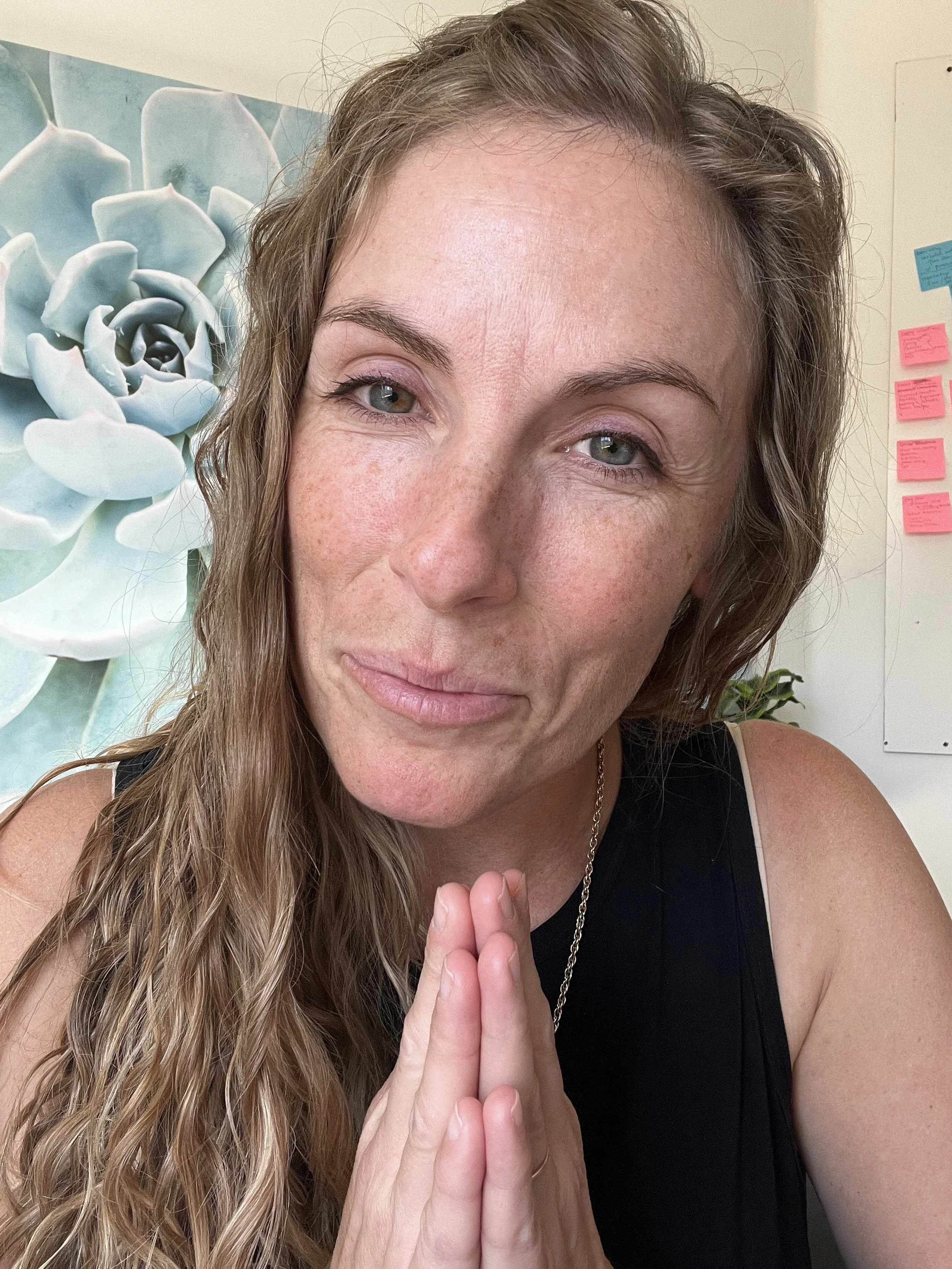 A woman with long, wavy hair and light skin, holding her hands in a prayer position, looking at the camera with a gentle smile, in an indoor setting with a succulent artwork and colorful sticky notes on the wall in the background.