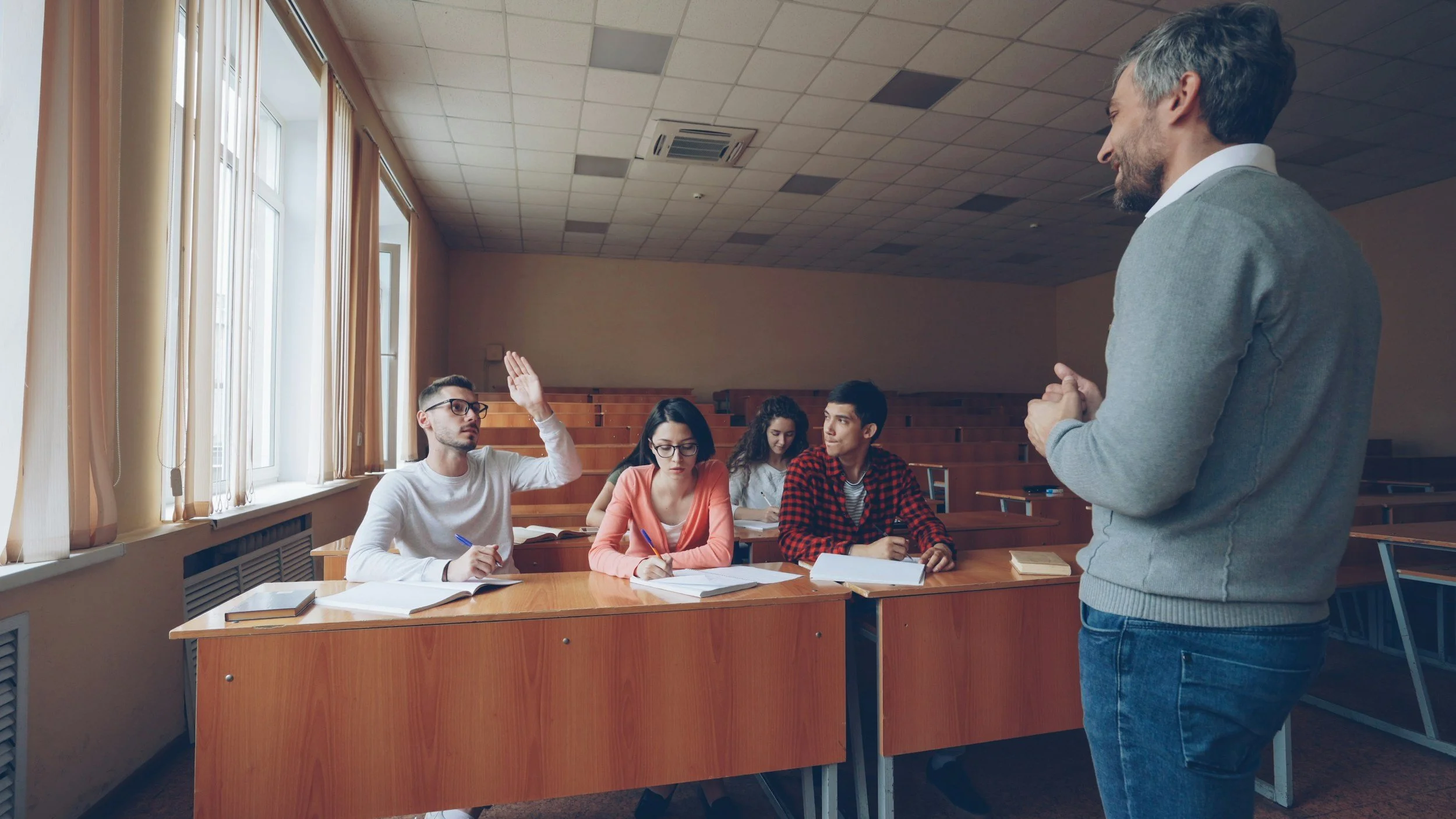A classroom scene with a teacher standing and four students sitting at desks, one student raising his hand. The classroom has large windows on the left, with beige curtains, and a tiled ceiling.