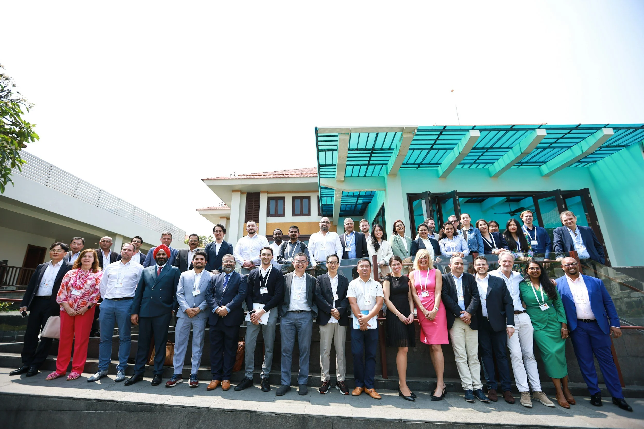 A large group of diverse education professionals in business attire posing for a photo outside a modern building with a blue roof canopy.
