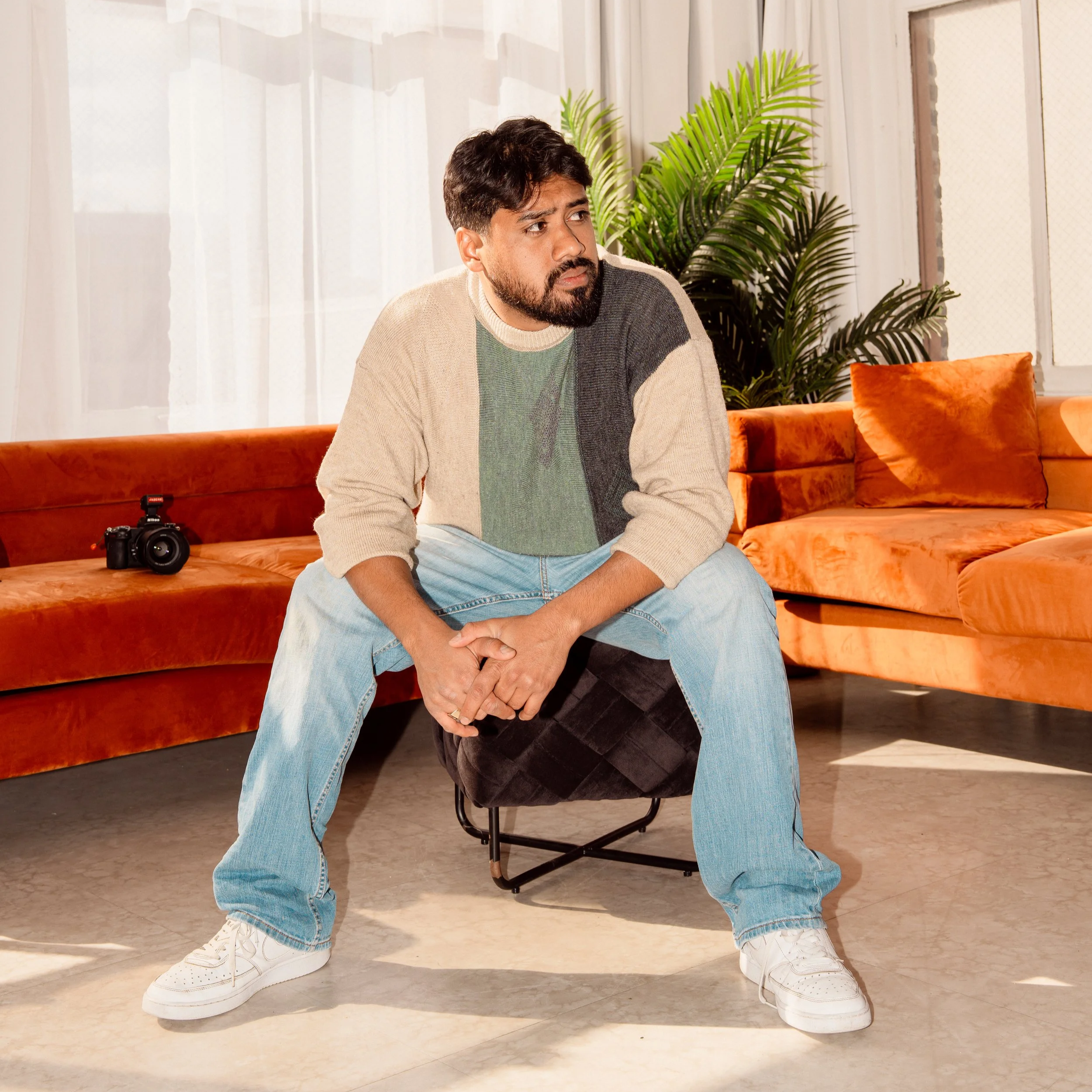 A young man sitting on a chair with his legs wide apart, leaning forward. He has medium-dark hair, a beard, and is wearing a tan and gray sweater, light blue jeans, and white sneakers. A camera is placed on the sofa beside him. The sofa is orange and the background features white curtains and green plants.