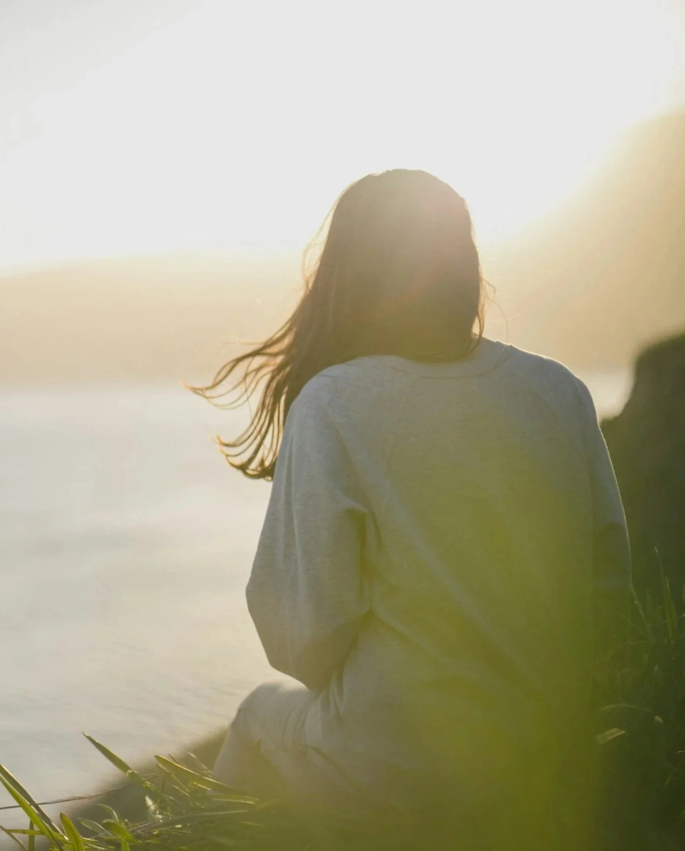 A woman with long hair sitting outdoors near water at sunset, facing away from the camera.