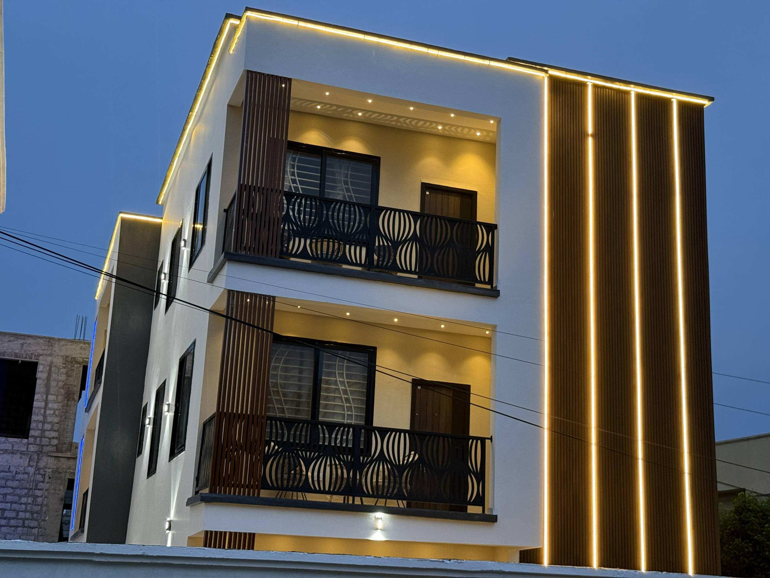 Modern multi-story building with illuminated vertical and horizontal LED lights, balconies with black decorative railings, and large windows night scene.